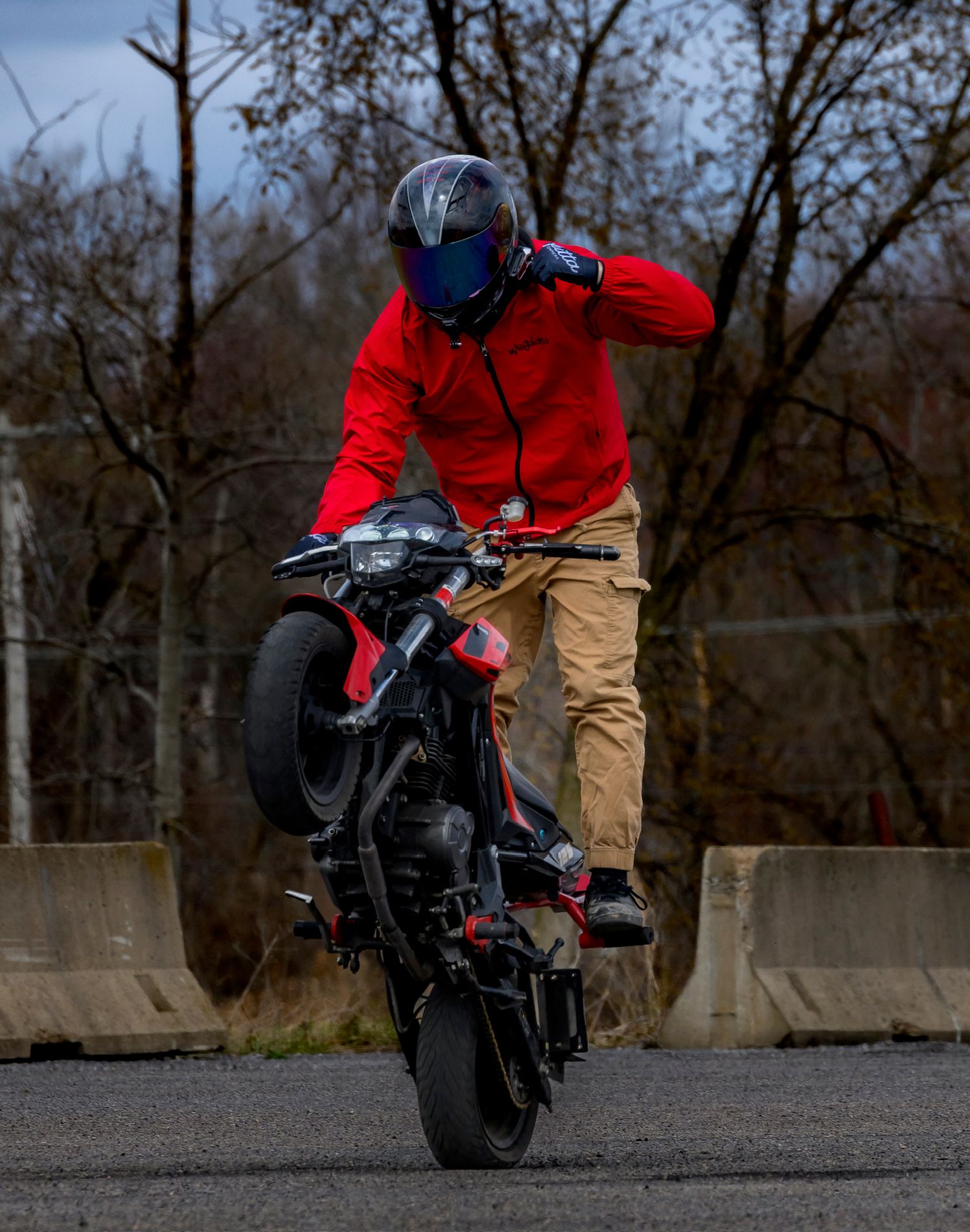 A stunt rider in a red hoodie and blue helmet performs a wheelie on a black motorcycle on an empty road.