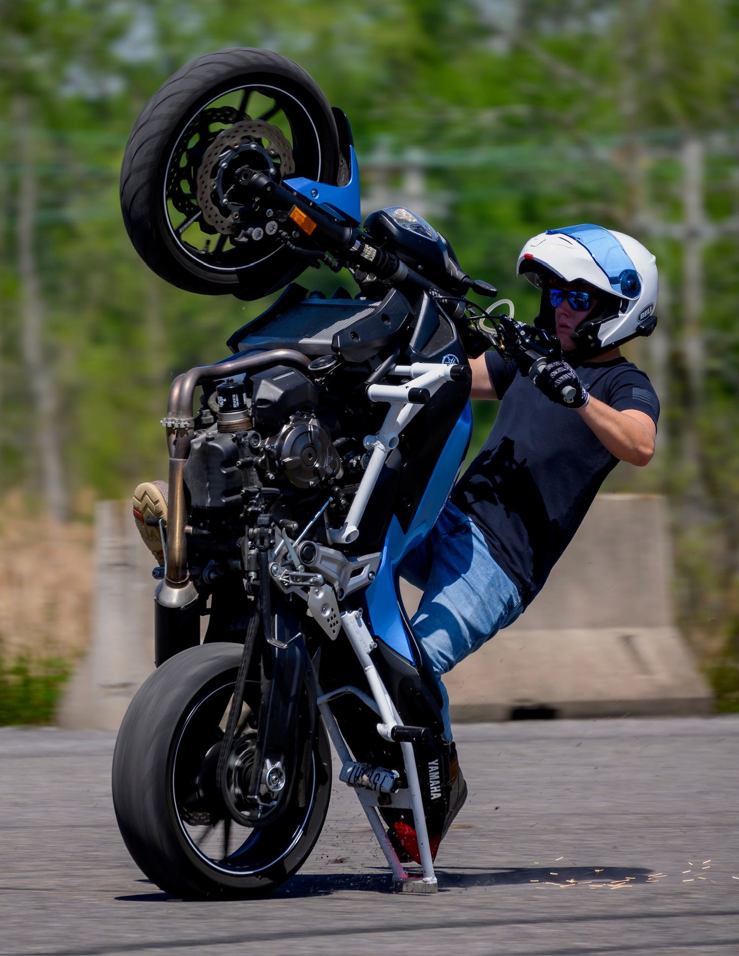 Stunt rider performing a wheelie on a blue motorcycle, wearing a helmet and protective gear on a paved surface.