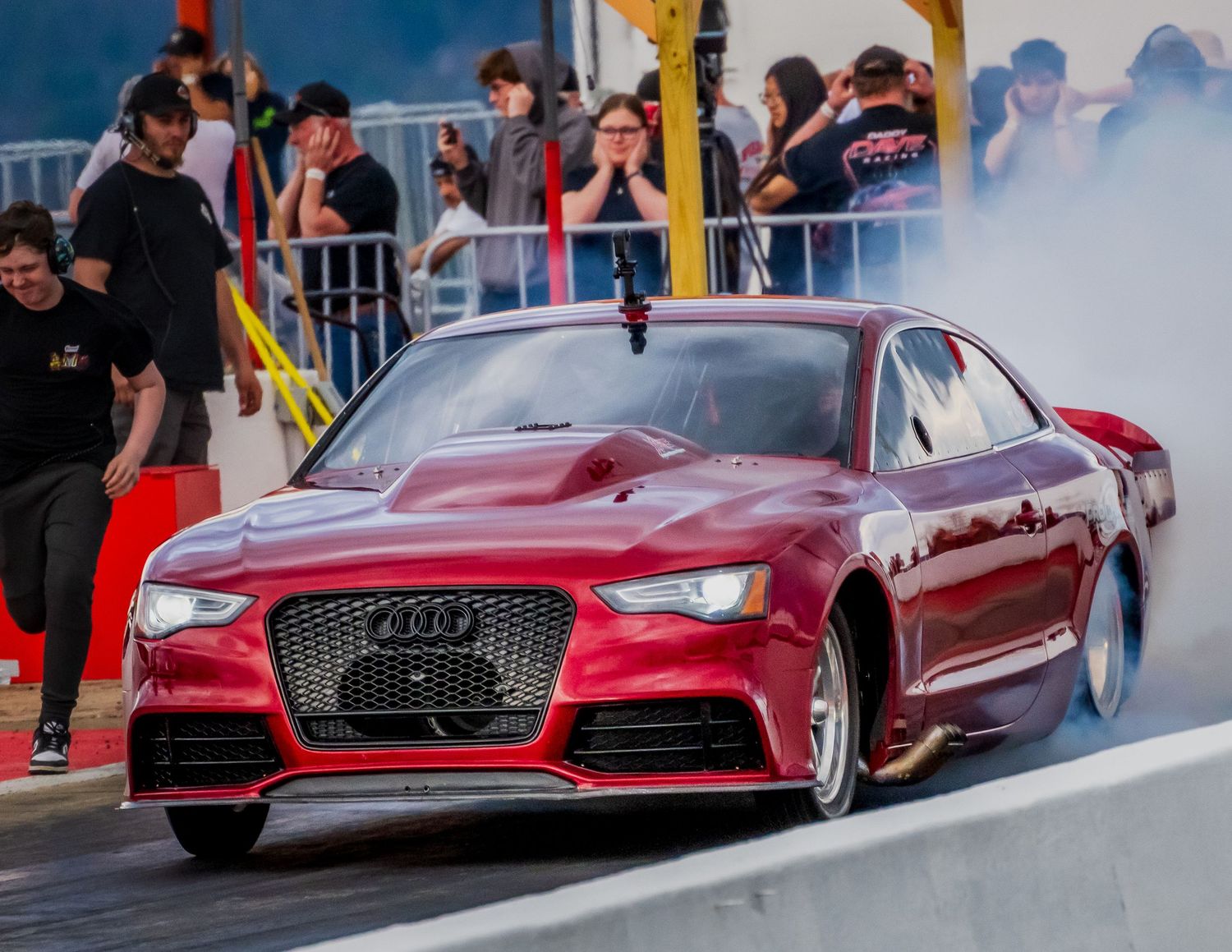 Red Audi sports cars lined up at a racing event with spectators watching.