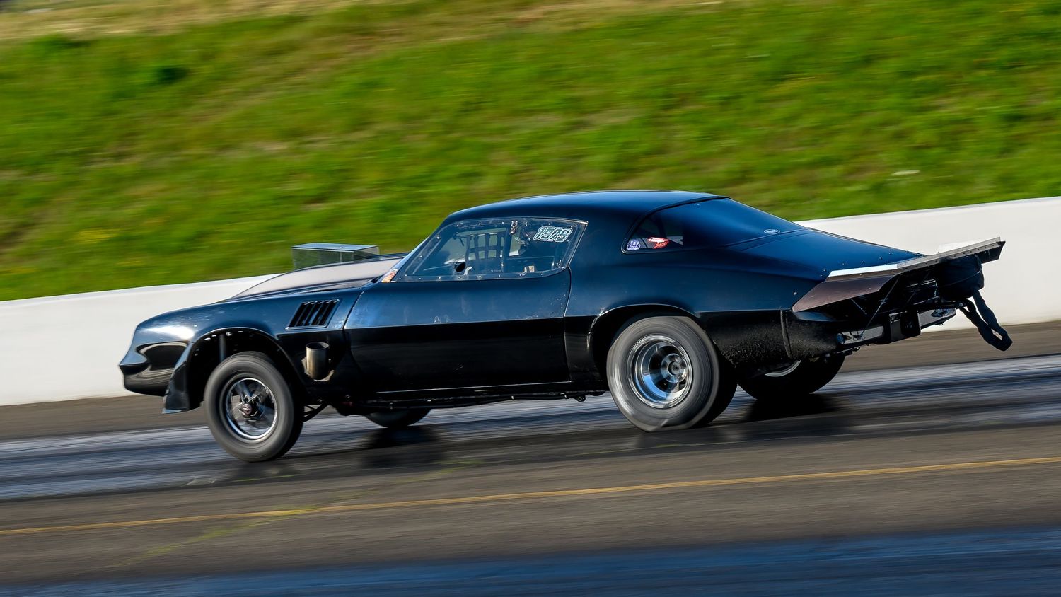 Drag racing cars speed past on a track with grass visible in the background.