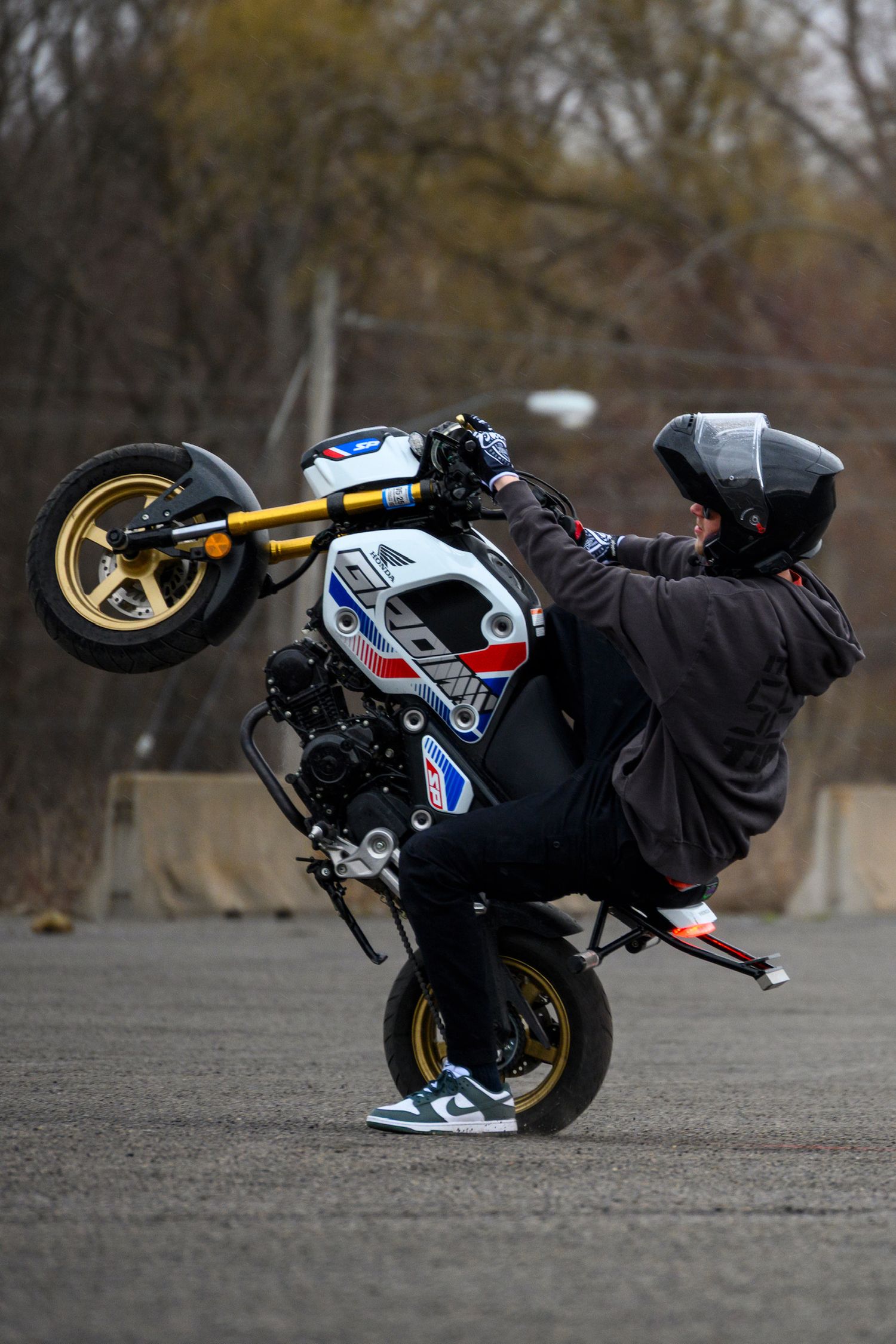 Stunt rider performing a wheelie on a mini motorcycle on an empty road wearing a helmet and dark clothing.