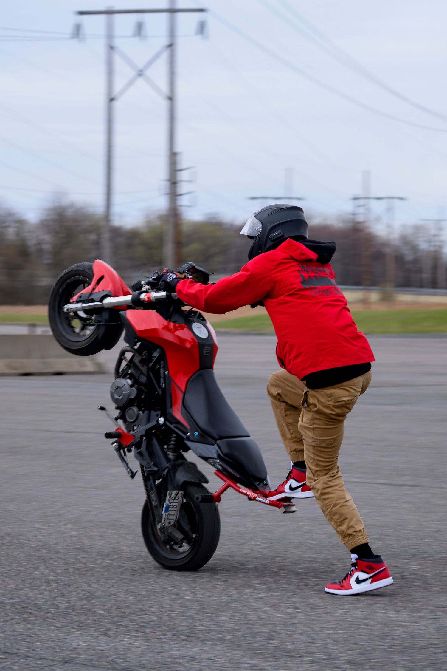 Stunt rider in red jacket and Nike Jordans performs a wheelie on a red motorcycle in an empty parking lot.