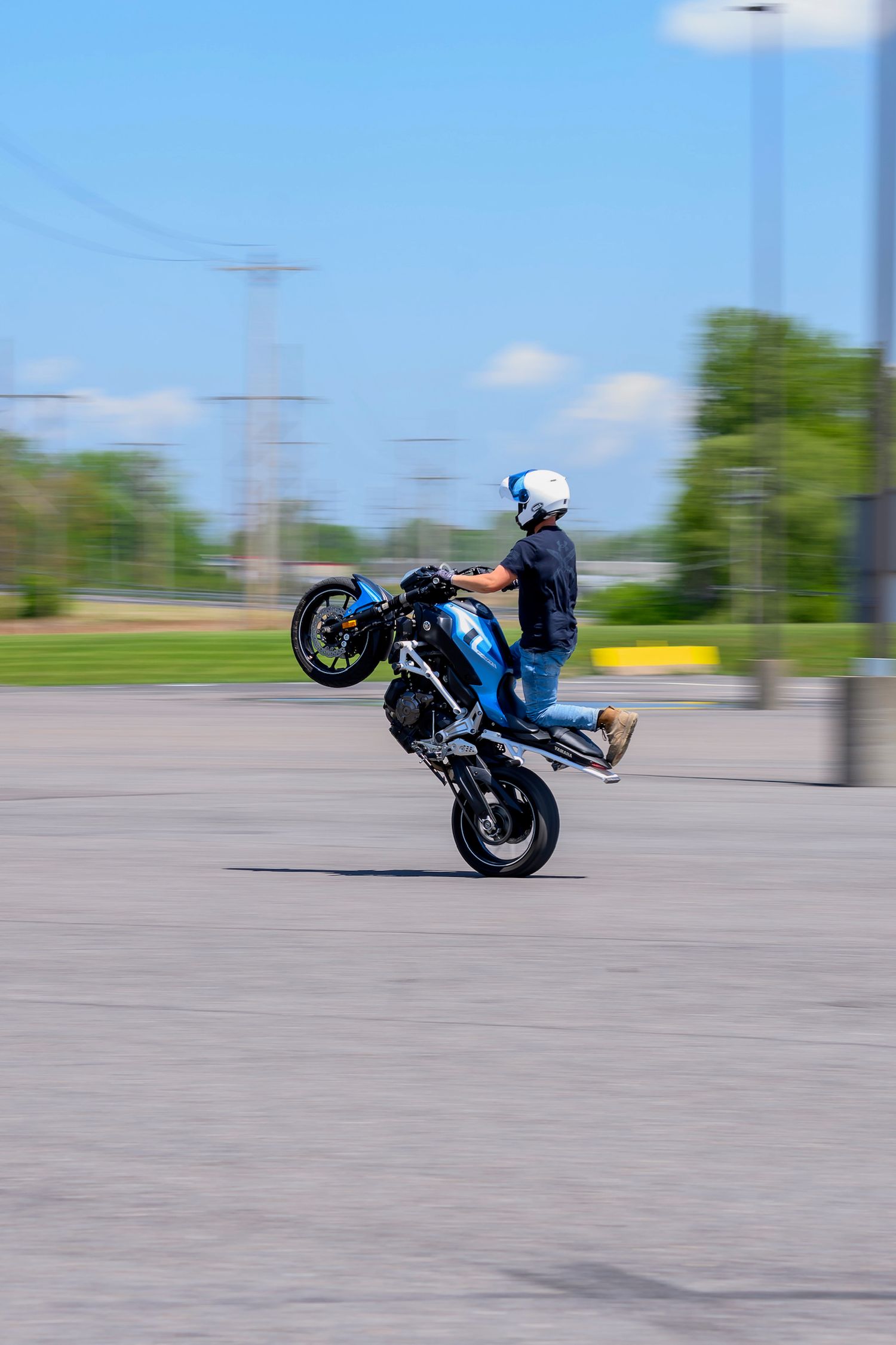 Stunt rider performing a wheelie on a blue motorcycle in an open parking lot under a clear blue sky.