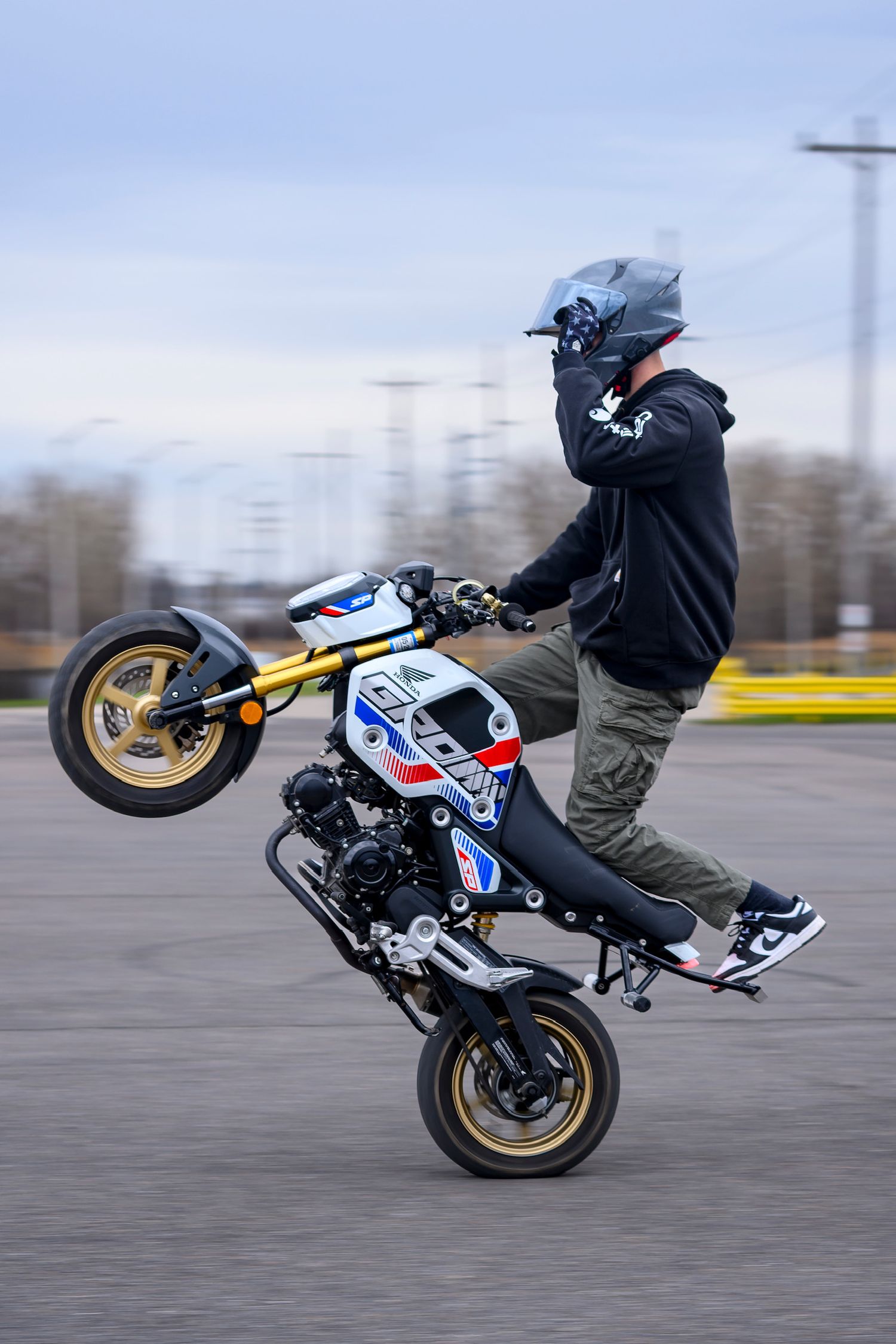 Stunt rider performing a wheelie on a Honda Grom mini motorcycle in a parking lot, wearing a helmet and black hoodie.