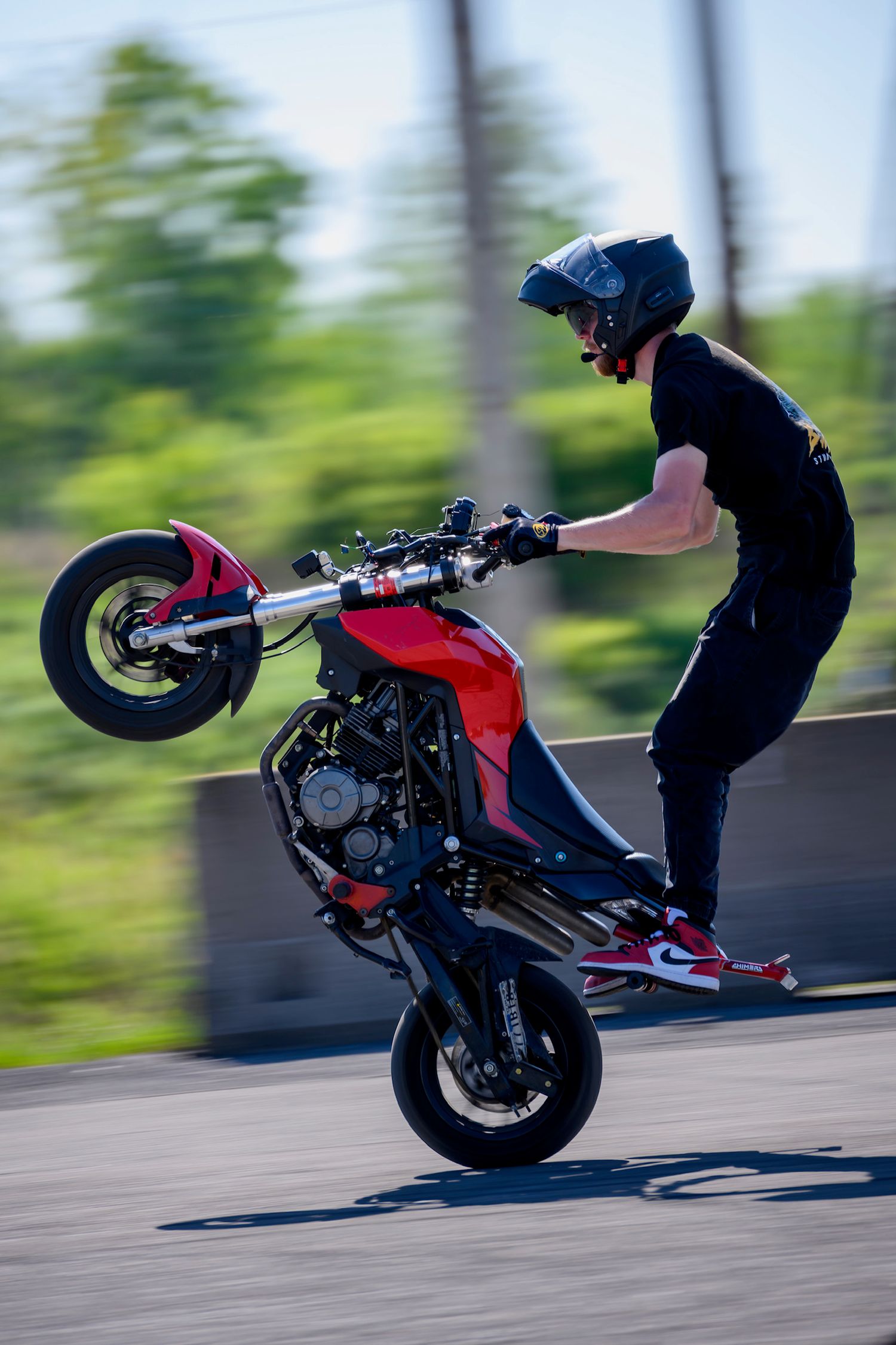 Motorcyclist performing a wheelie stunt on a red bike while standing, wearing black gear and helmet on a road.