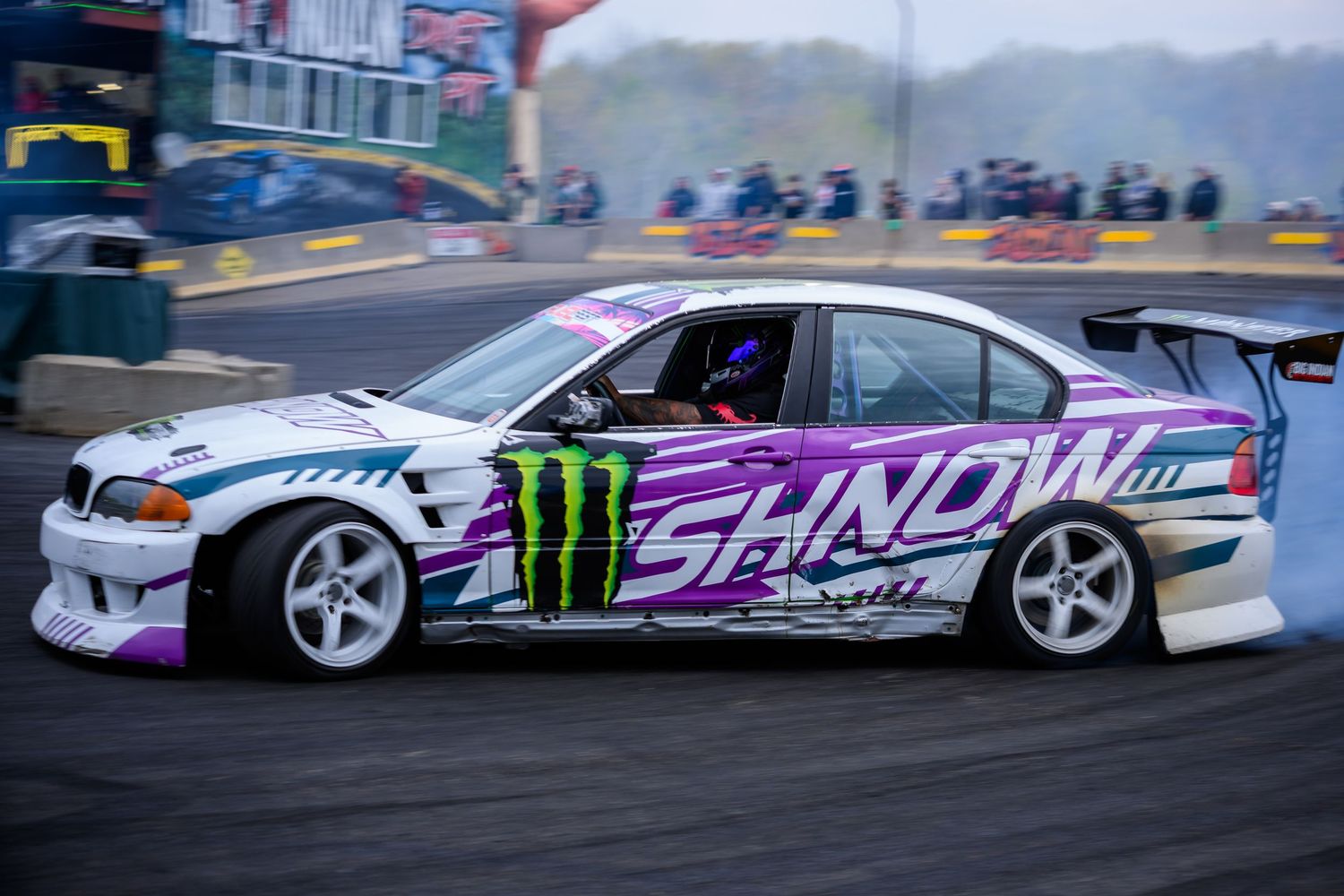 Purple and white SHNOW Monster Energy drift car racing on a track, performing a sideways drift maneuver.