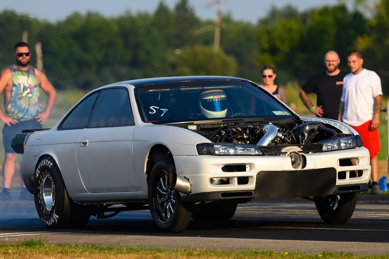 Multiple sports cars lined up at a racing event with spectators watching from the sidelines.