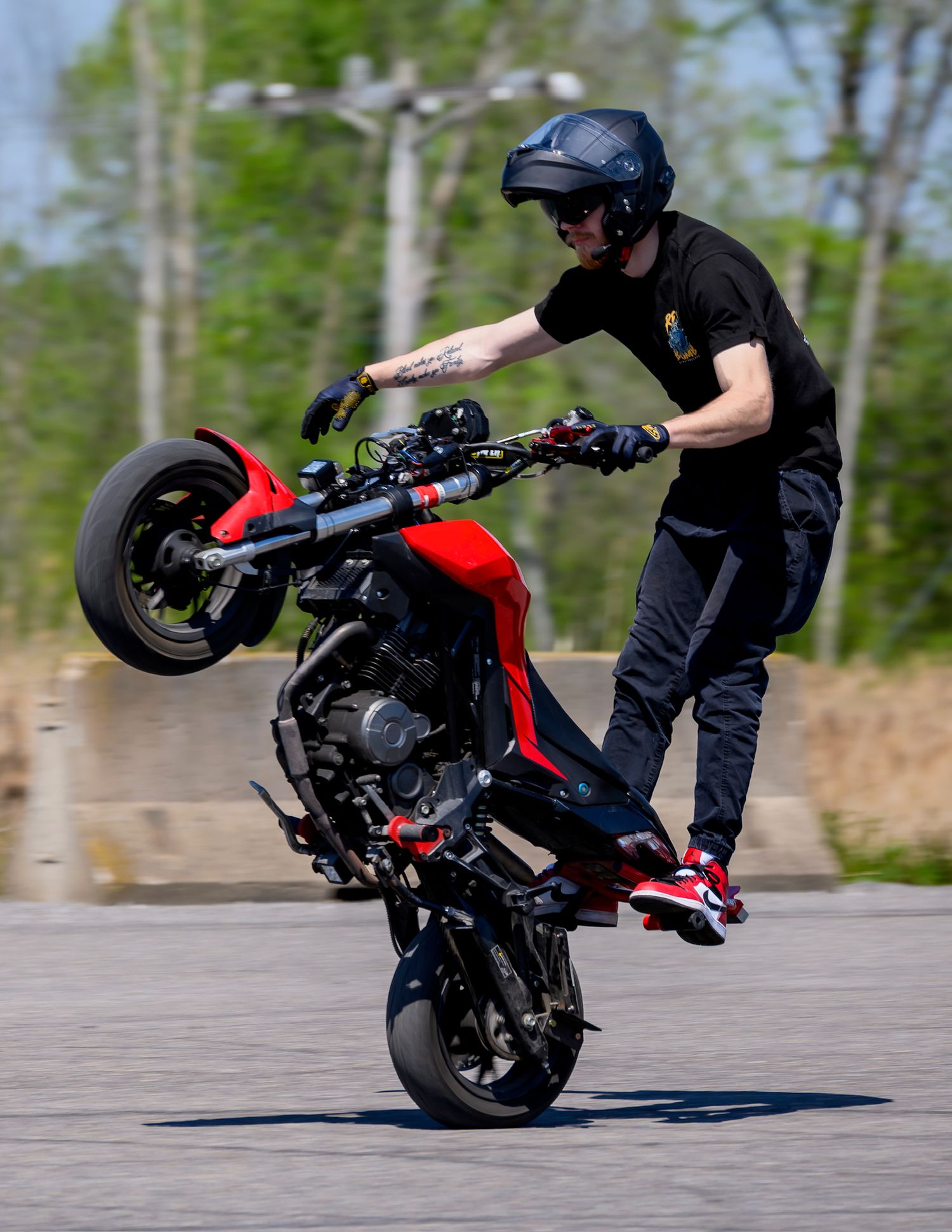 A stunt rider performs a wheelie on a red motorcycle in a parking lot, wearing a blue helmet and black outfit.
