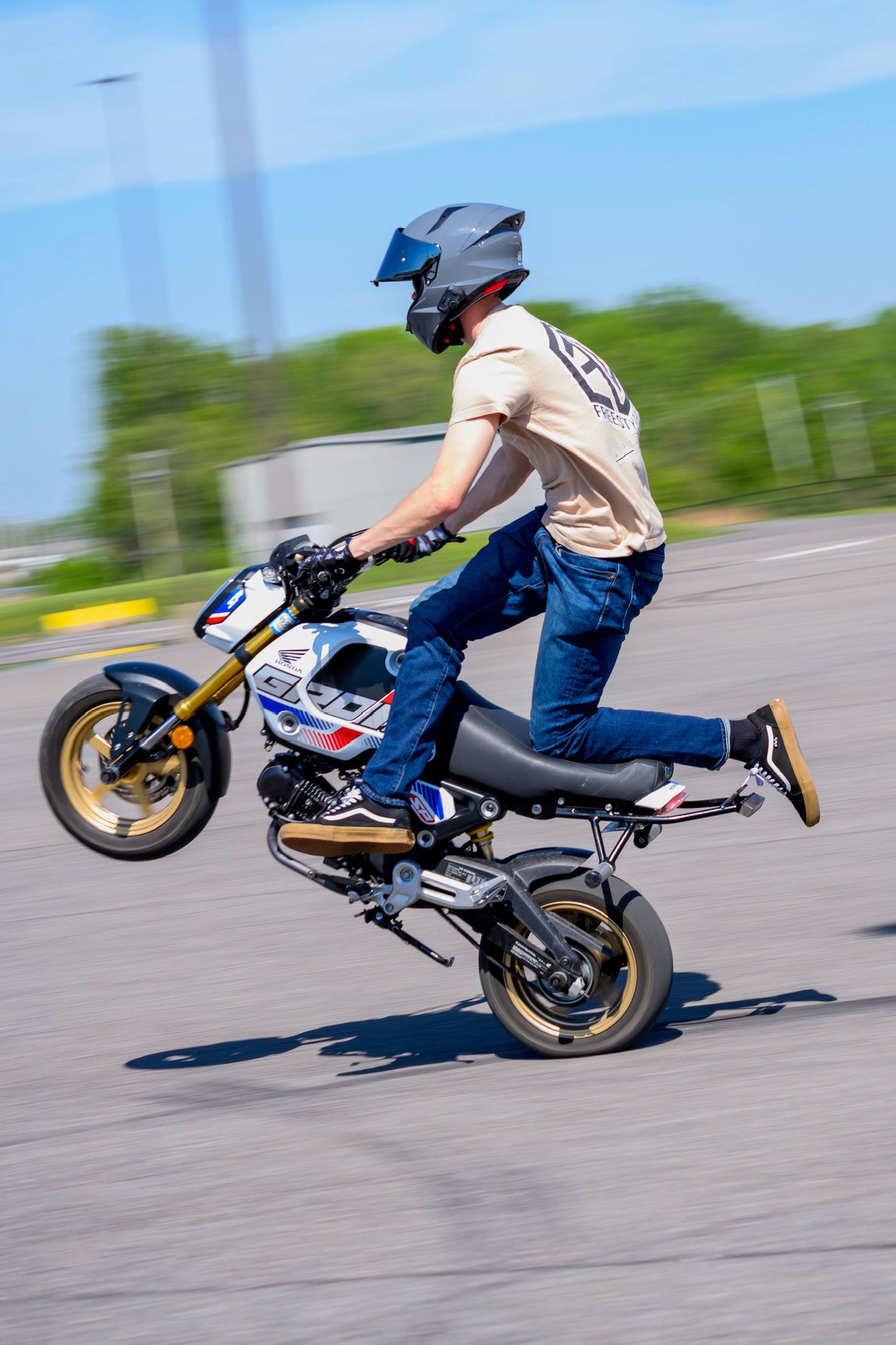 Motorcyclist performing a stunt wheelie on a small sport bike while wearing a helmet and jeans on an open pavement area.