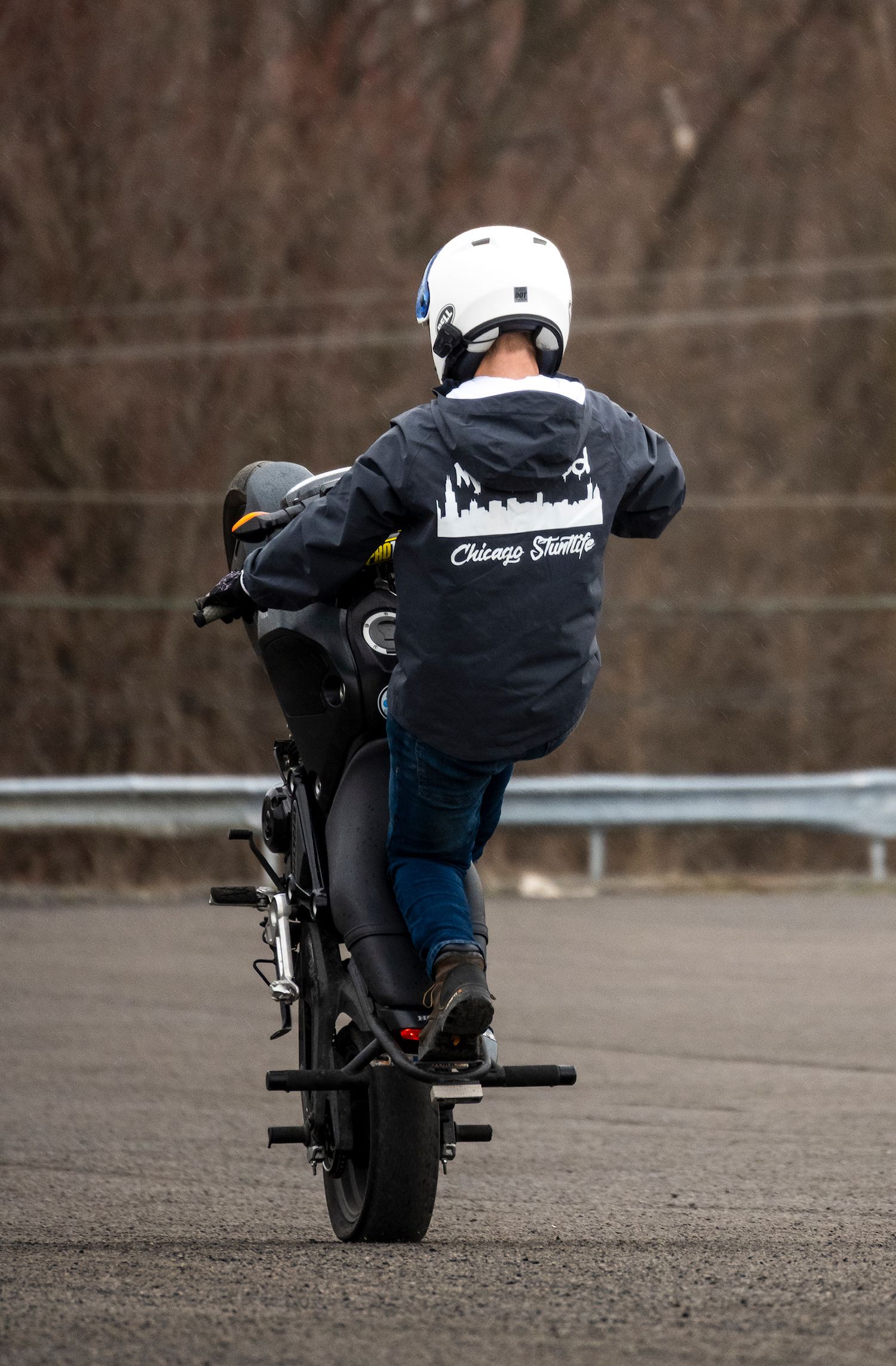 Motorcycle stunt rider performing a wheelie on a black bike, wearing a white helmet and Chicago Streetlife jacket.