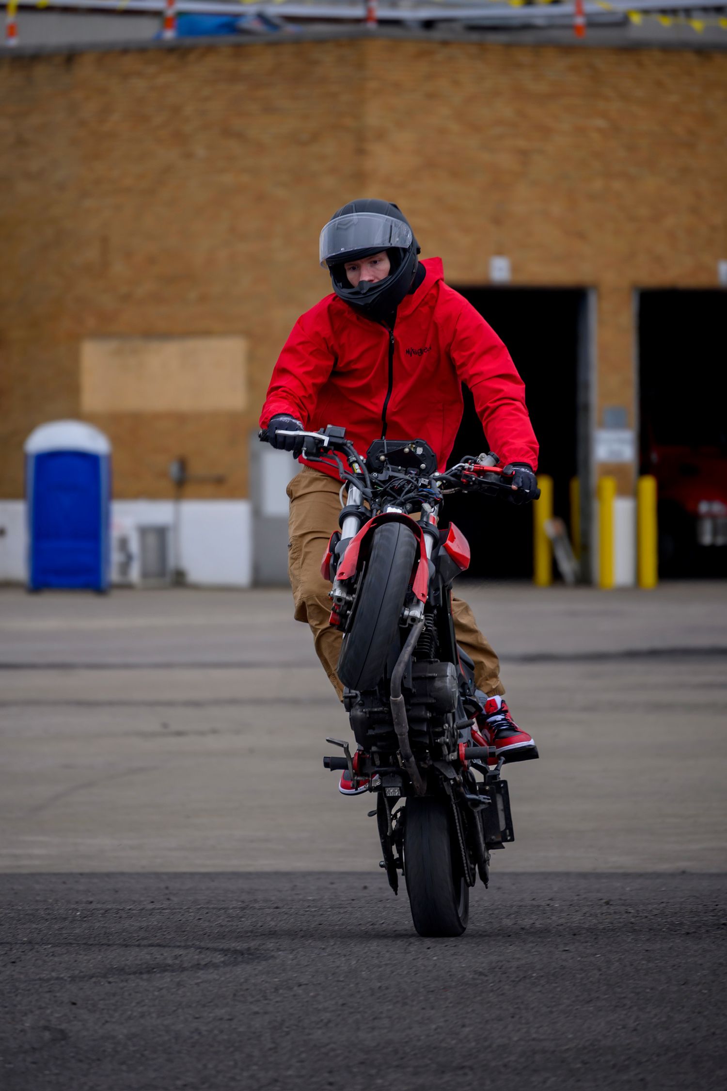 A motorcyclist in a red jacket and helmet performs a wheelie stunt in a parking lot near an industrial building.