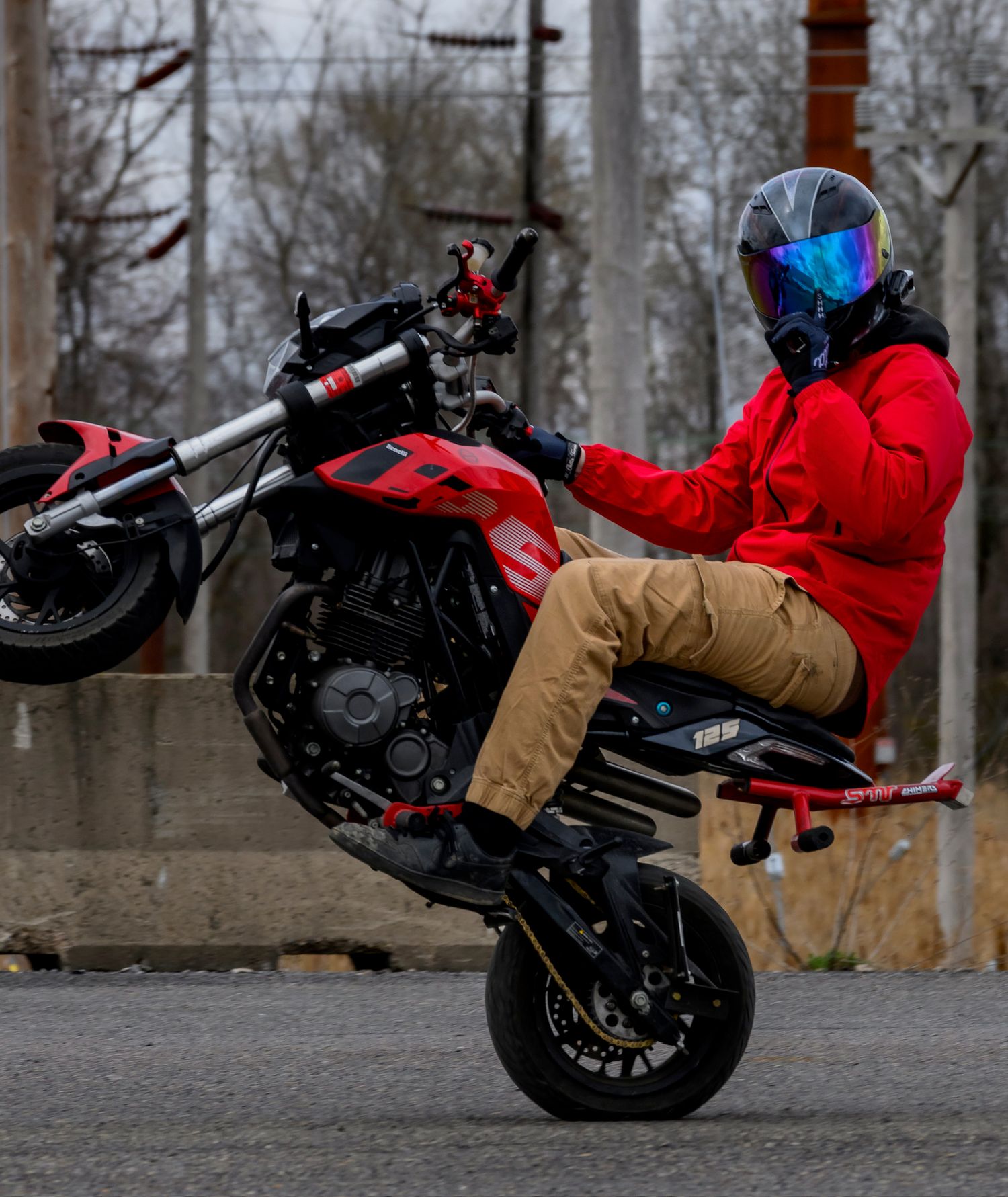 Motorcyclist in red jacket and blue visor helmet performing a wheelie stunt on a red sport bike on a street.