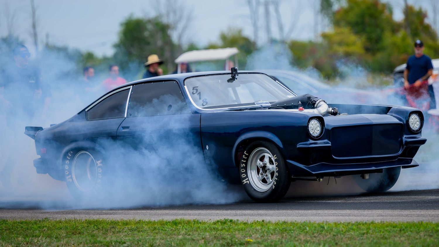 Vintage muscle cars performing burnouts and creating smoke clouds at a drag strip.