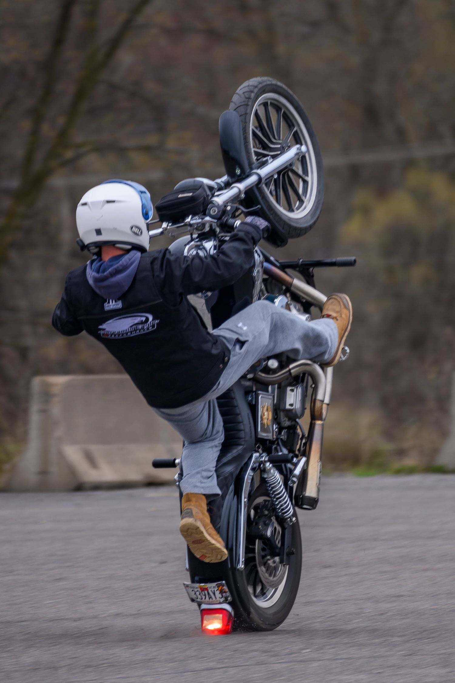 Motorcyclist performing a wheelie stunt on a street bike, wearing a white helmet and black jacket on a paved surface.
