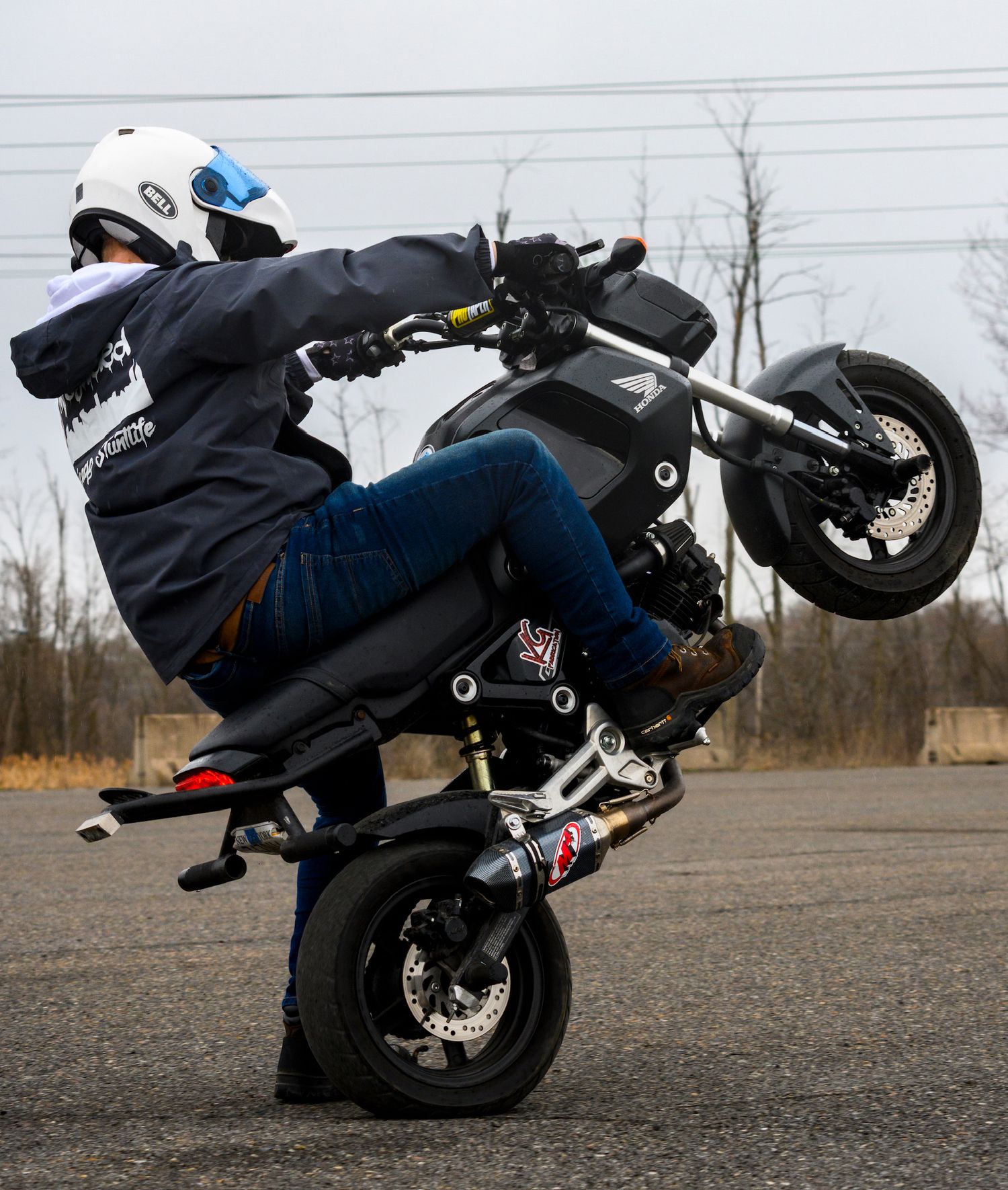 Motorcyclist performing a wheelie stunt on a small sport bike while wearing a white helmet and blue jeans.
