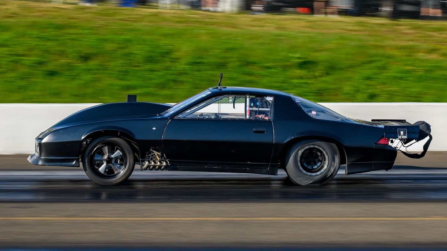 Dark colored drag racing cars line up on a racing track against a blurred background.