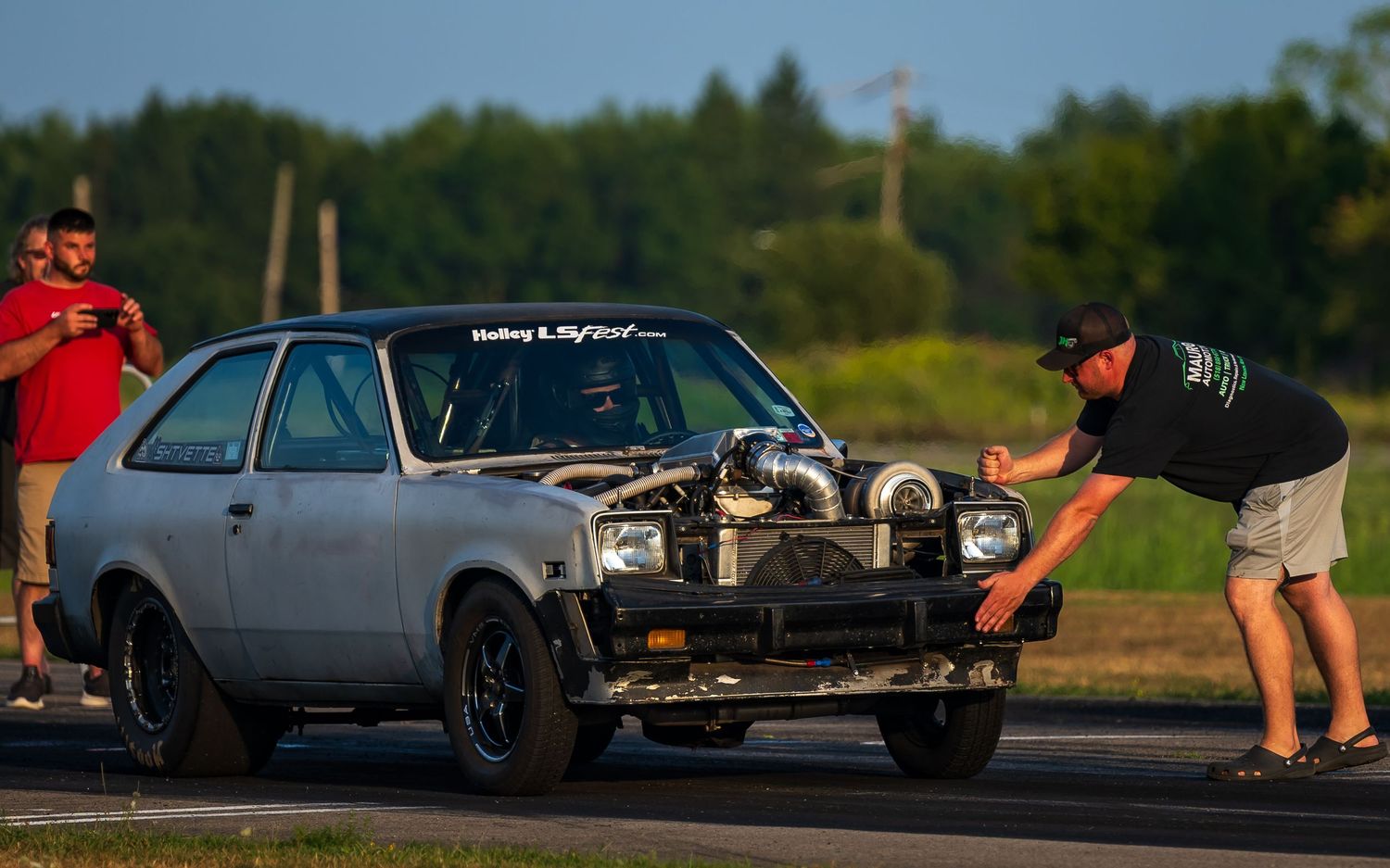Custom cars lined up at a motorsport event with spectators watching from the sidelines.