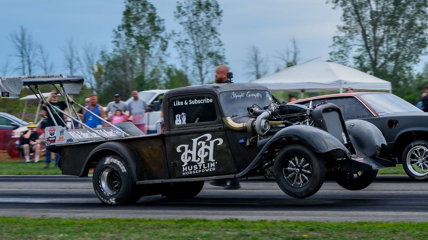 Custom racing vehicles lined up at a drag racing event with spectators watching.