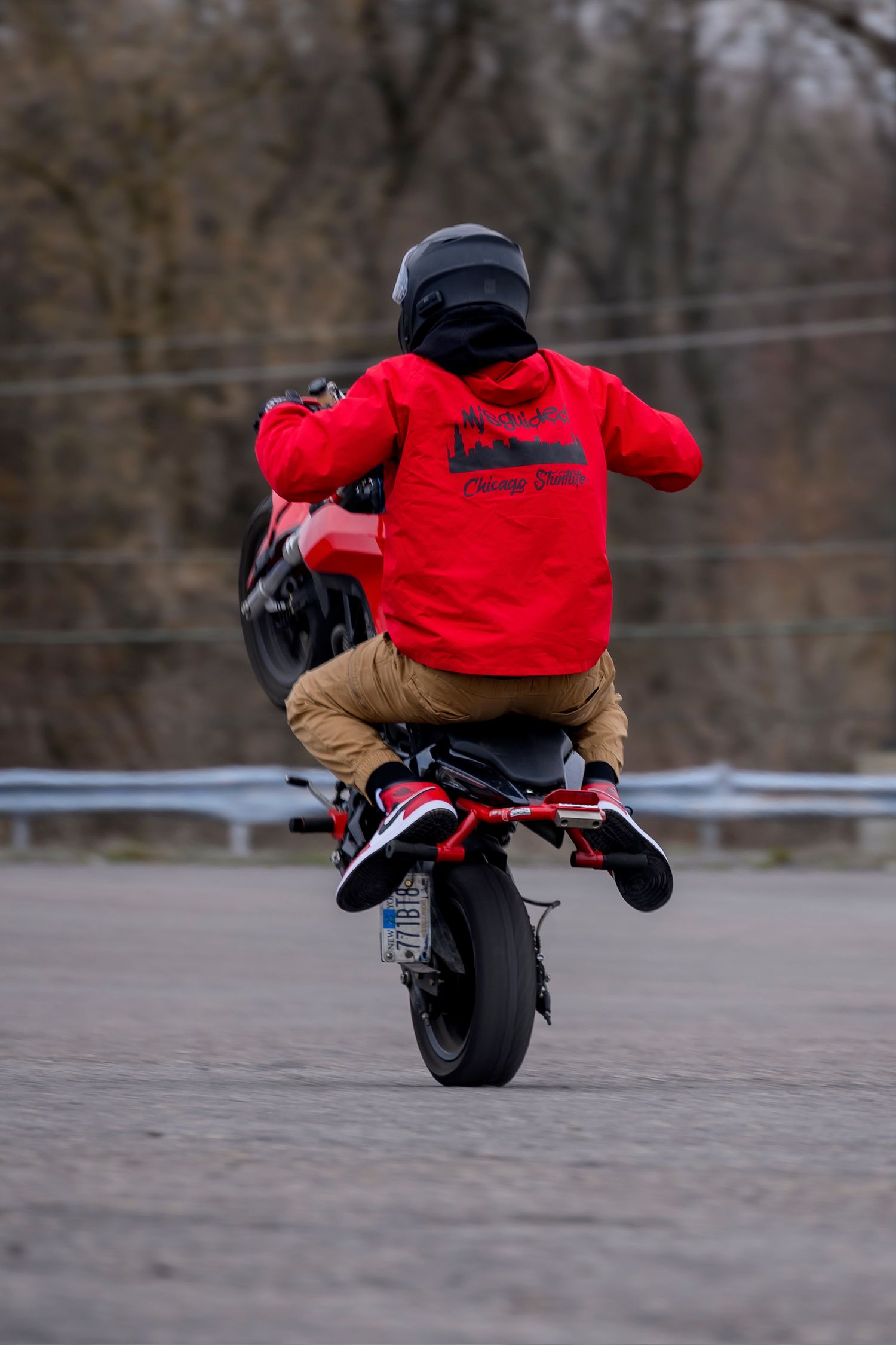 Motorcyclist in red jacket performs a wheelie stunt on a red bike, wearing helmet and sneakers on a paved road.