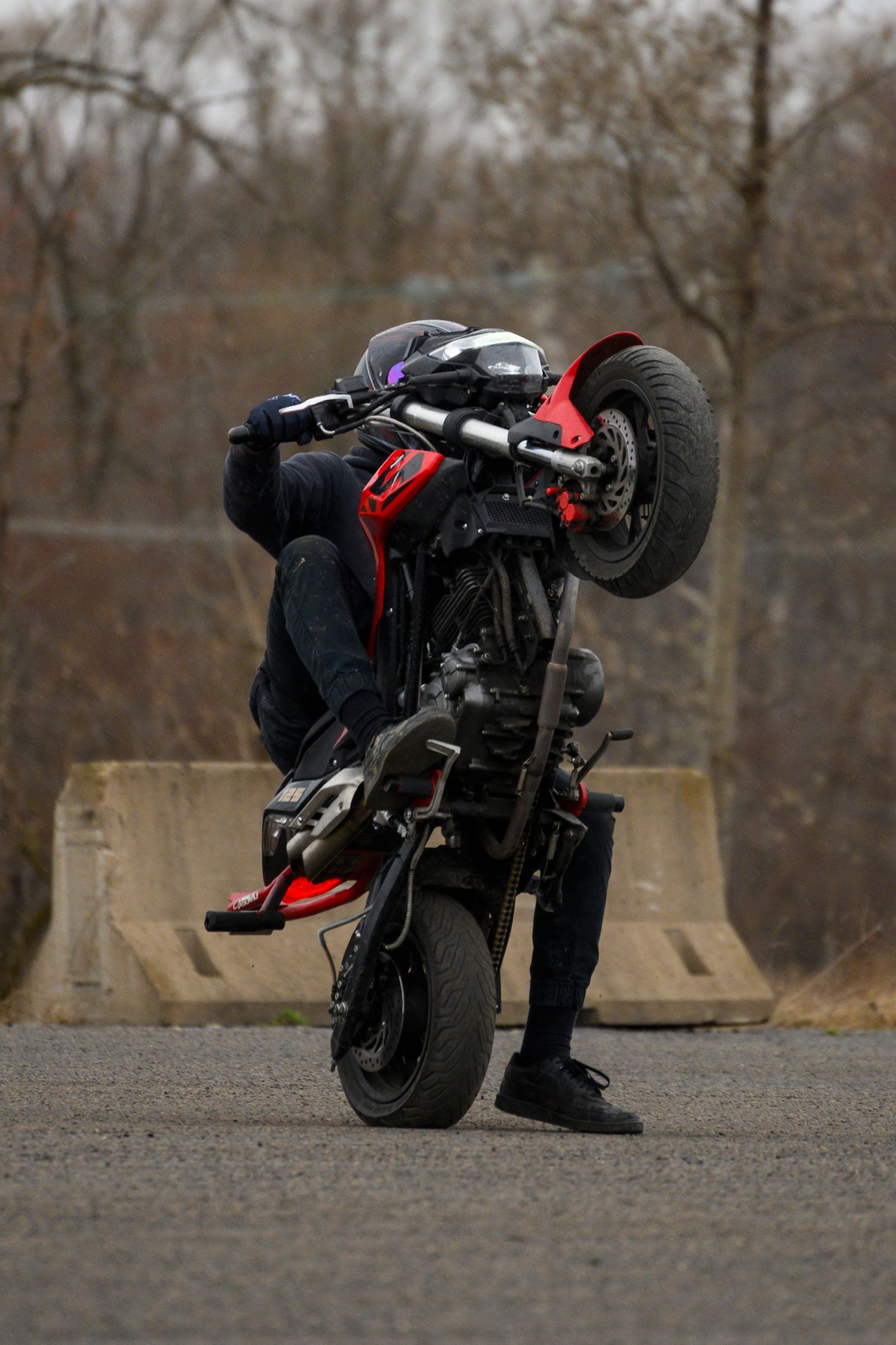 A motorcyclist performs a wheelie stunt on a red and black bike on an empty road with bare trees in the background.