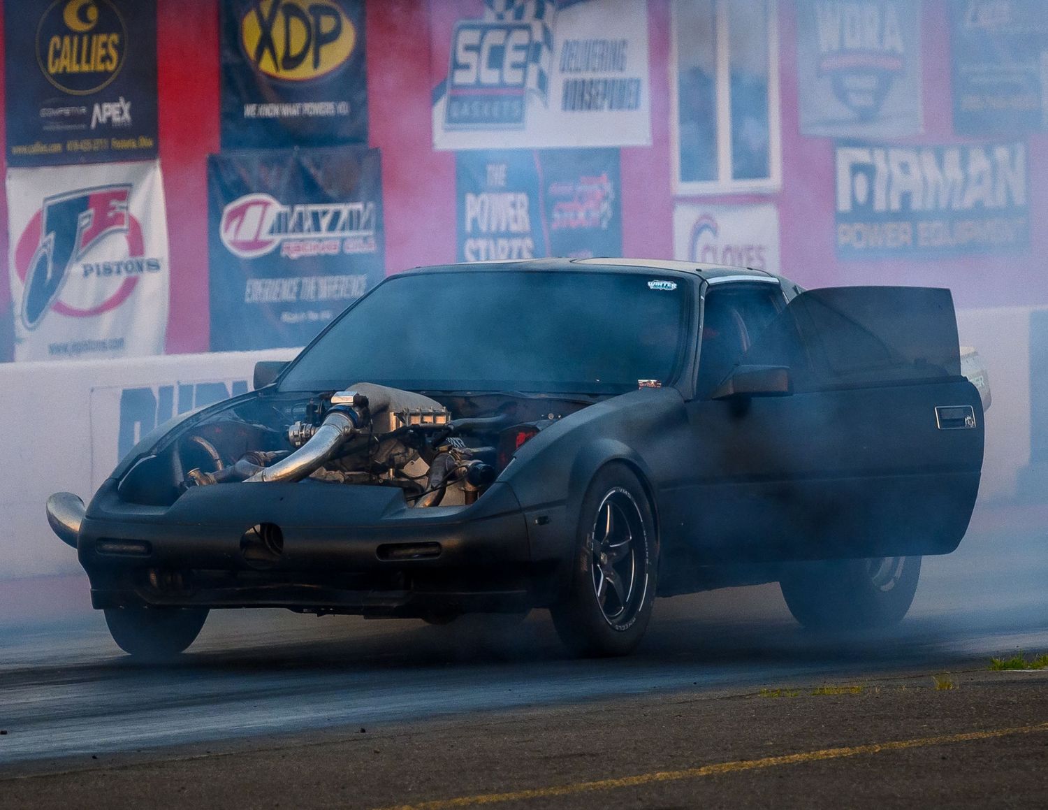 A drag racing car creates smoke during a burnout with sponsor banners visible in the background.