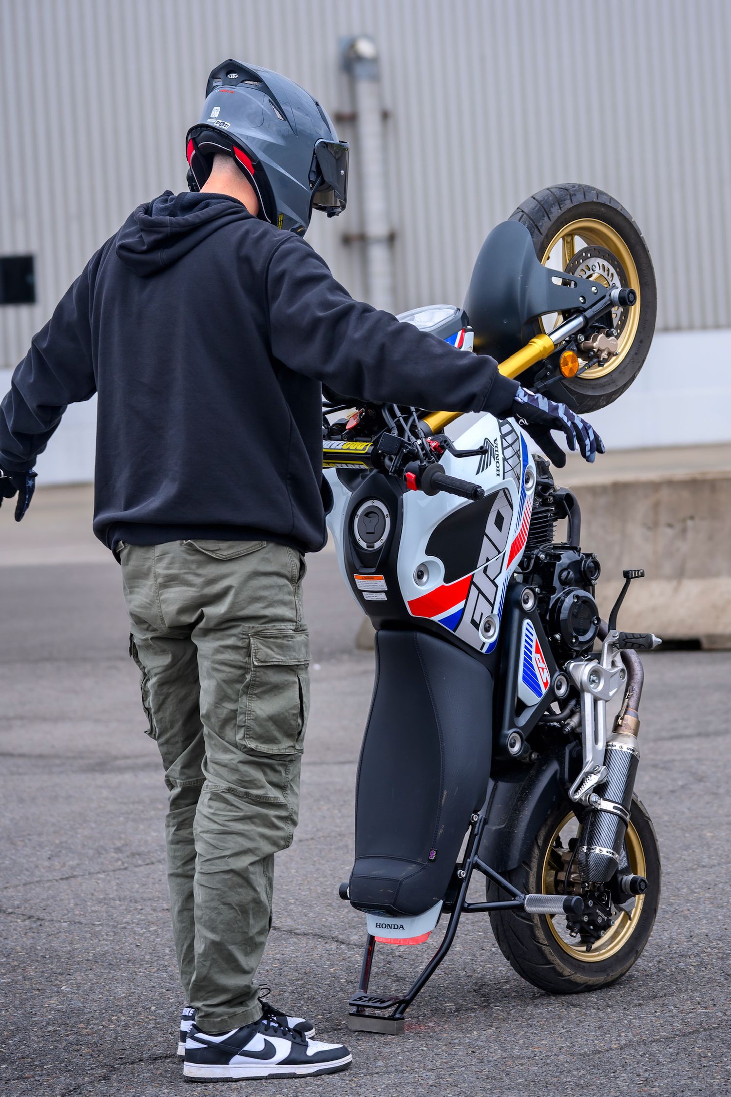 Person in helmet and cargo pants performing a wheelie stunt on a small sport motorcycle in a parking lot.
