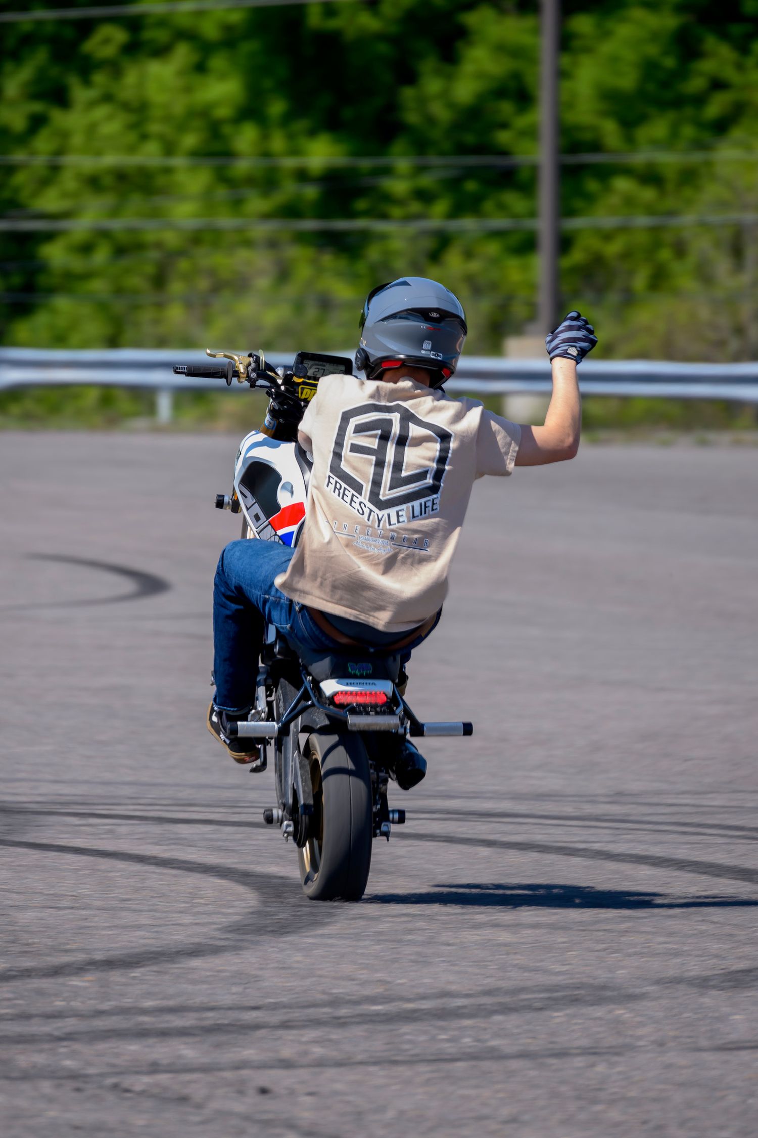 Young rider performing a wheelie on a small motorcycle in a parking lot, wearing a helmet and Freestyle Life t-shirt.