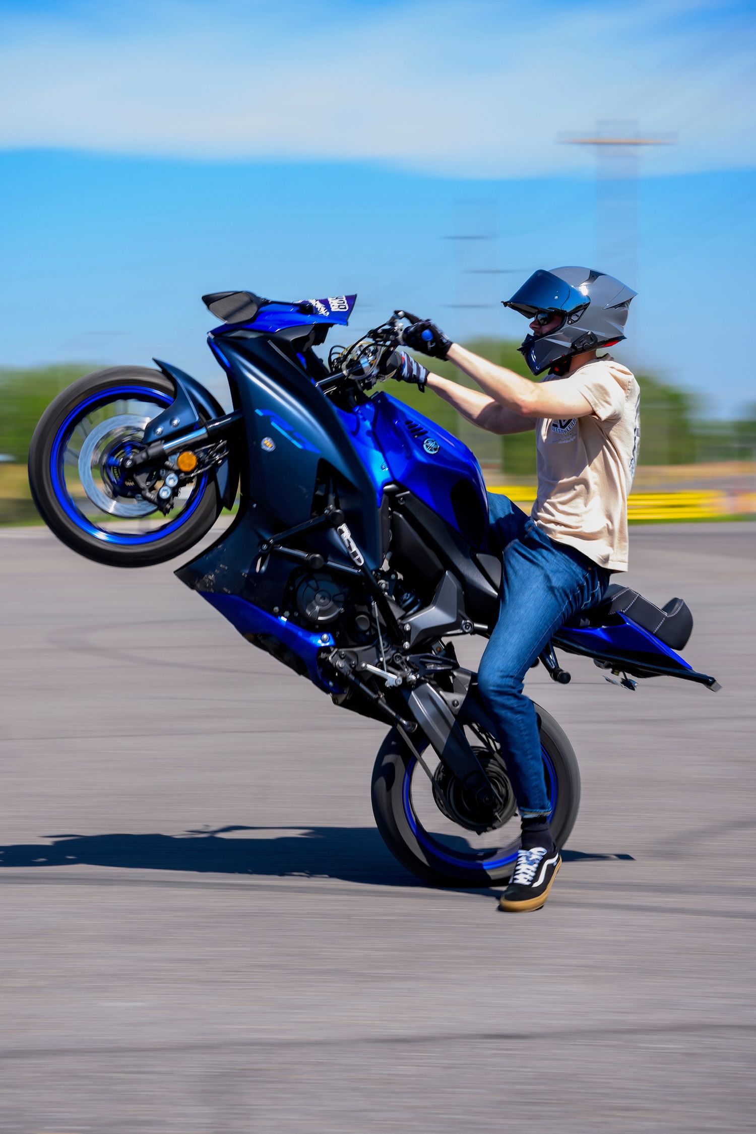 Motorcyclist performing a wheelie stunt on a blue sport bike while wearing a helmet and jeans on an open road.