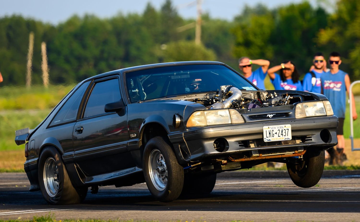 A black modified Mustang with a large engine protruding from the hood on a drag strip.