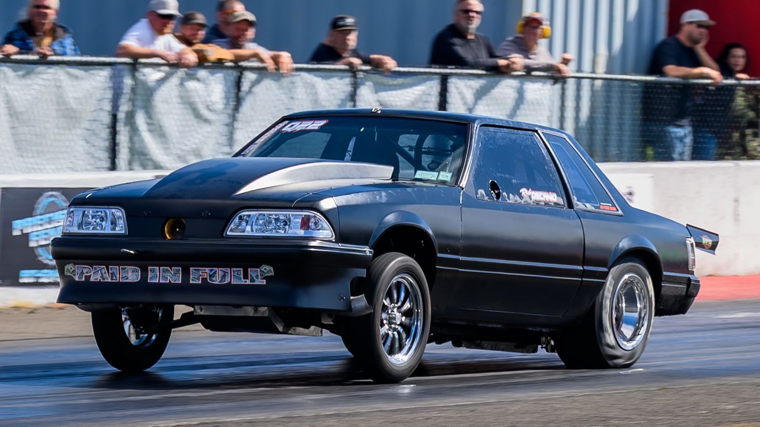Performance cars lined up for drag racing with spectators watching from behind barriers.