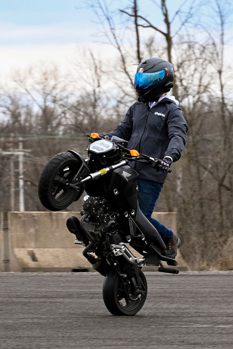 Motorcyclist performing a wheelie stunt on a black sport bike while wearing a helmet and riding gear outdoors.