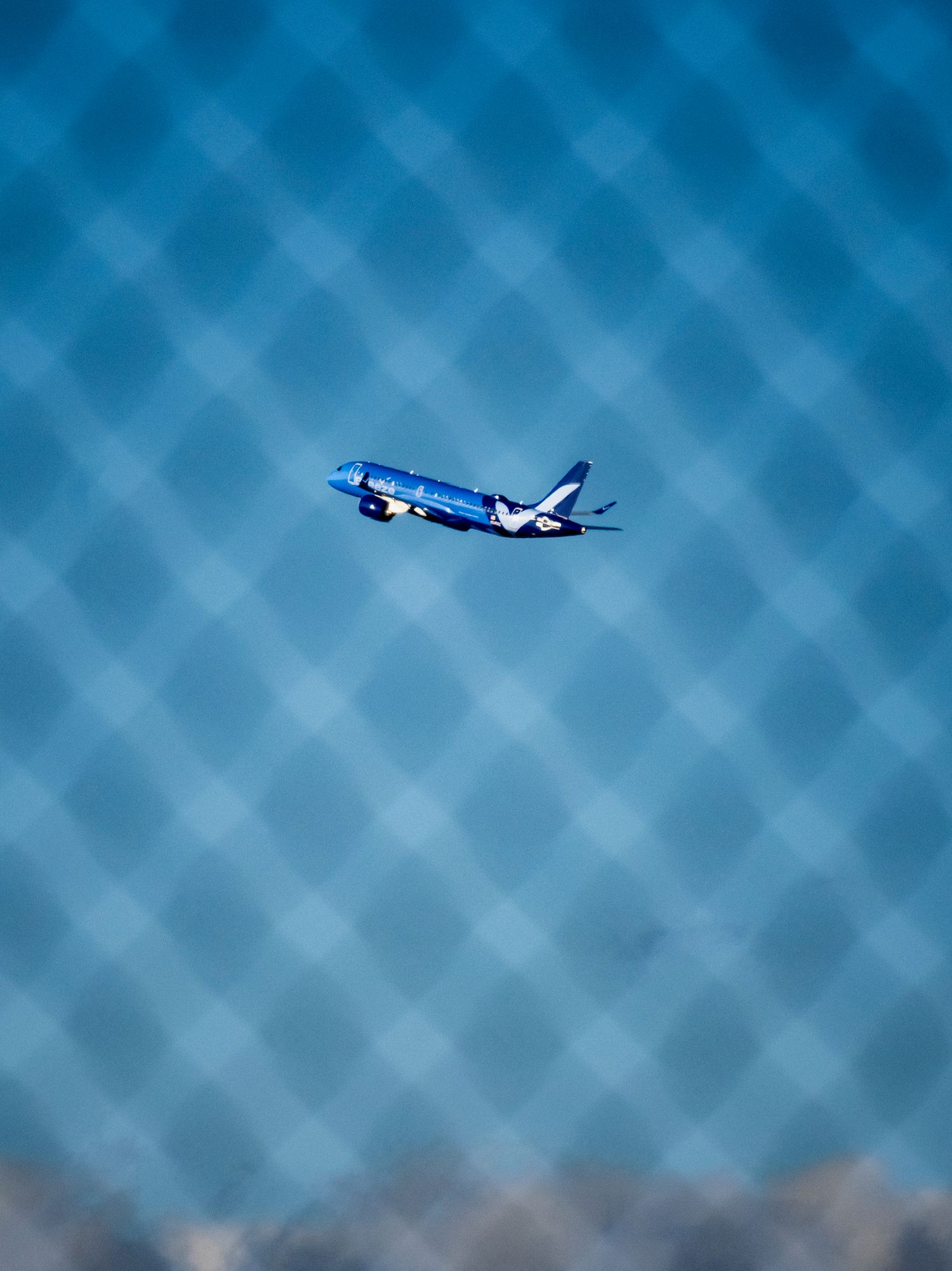 Blue commercial airplane flying through clear blue sky, photographed through a diamond-patterned fence or mesh.