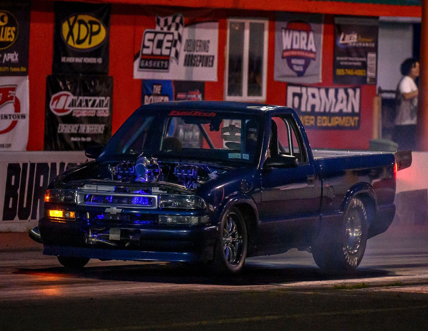 A dark drag racing car performs a burnout at night with red illuminated signage in the background.