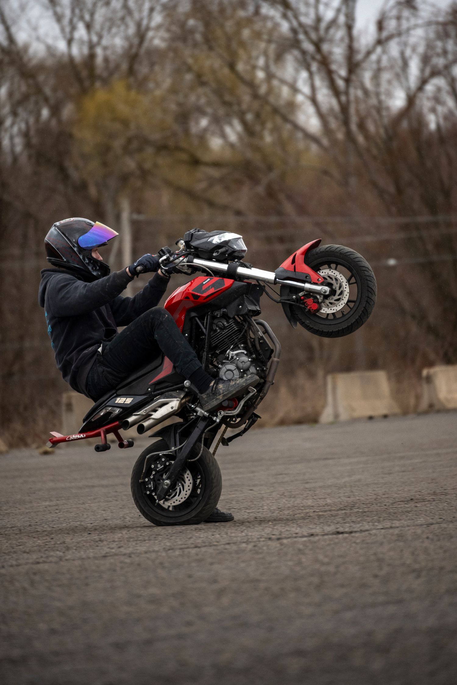 Motorcyclist performing a wheelie stunt on a red supermoto bike in a parking lot wearing a helmet.