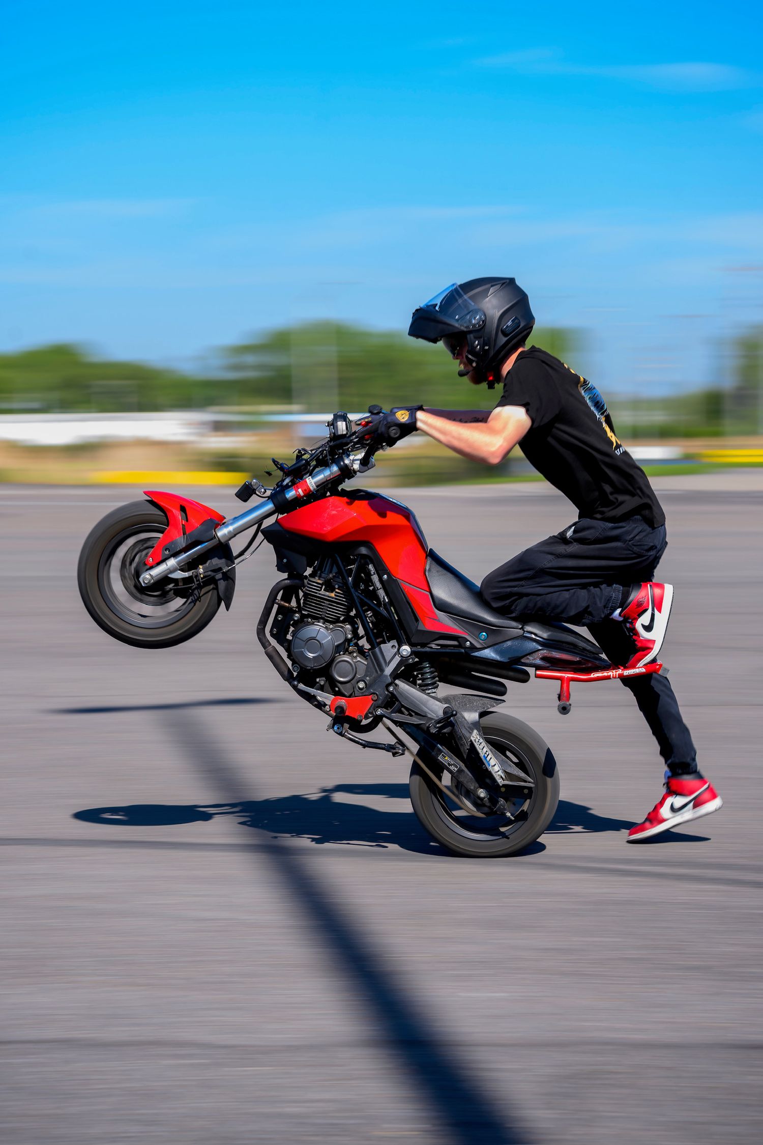 Stunt rider performing a wheelie on a red motorcycle wearing black outfit, helmet, and red Nike sneakers on a sunny day.