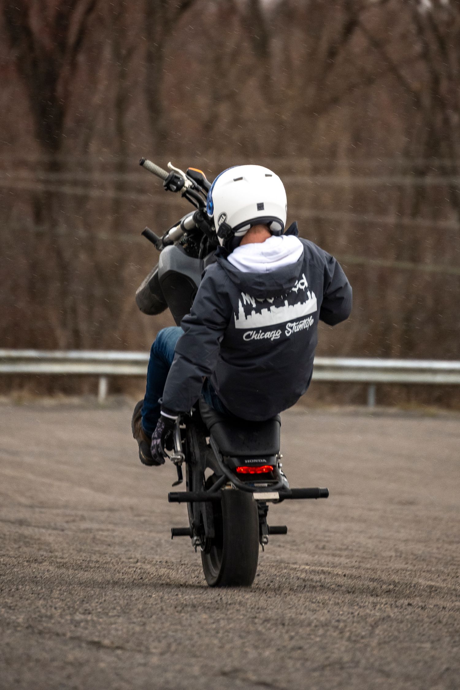 Motorcyclist performing a wheelie stunt on a parking lot, wearing a white helmet and Chicago Stunts jacket.
