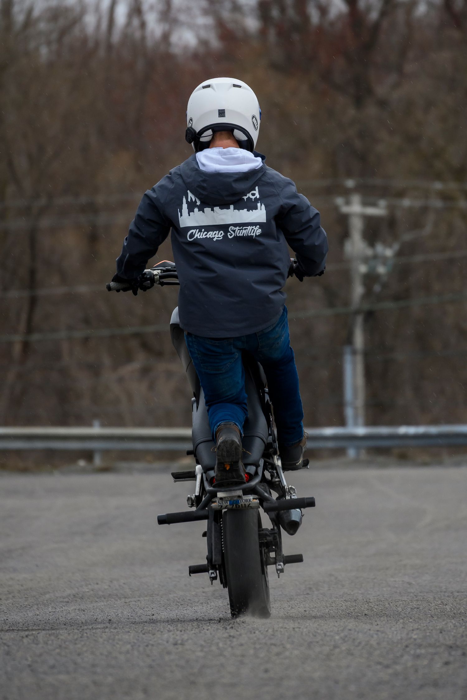 Motorcyclist wearing a white helmet and Chicago Shuttle jacket performs a wheelie on a dark sport bike on an empty road.