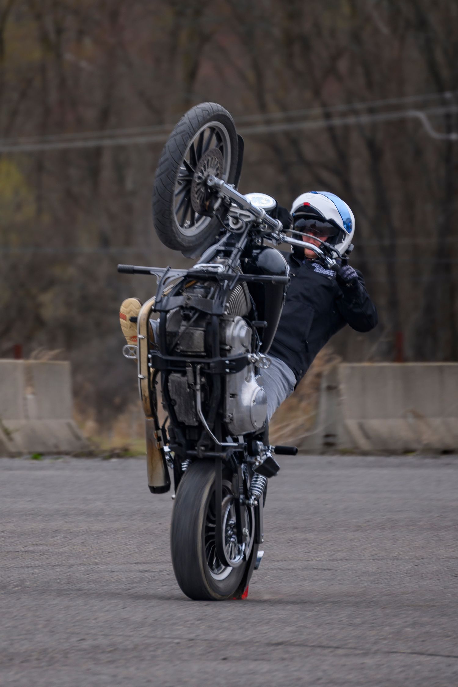 Motorcyclist performing a wheelie stunt on a classic motorcycle in a parking lot, leaning back dramatically.