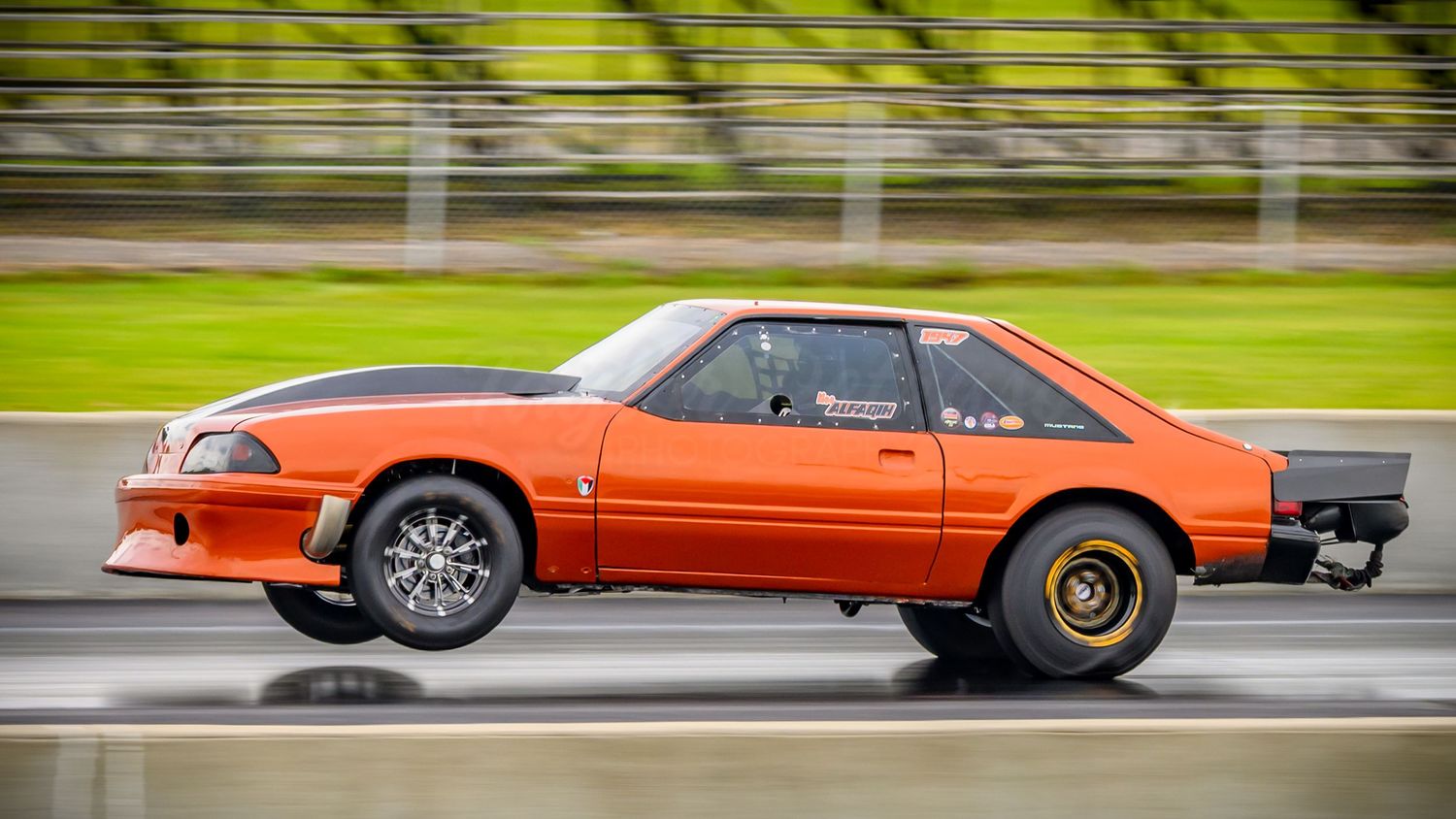 Classic orange muscle cars lined up at a motorsports event on a green racetrack.