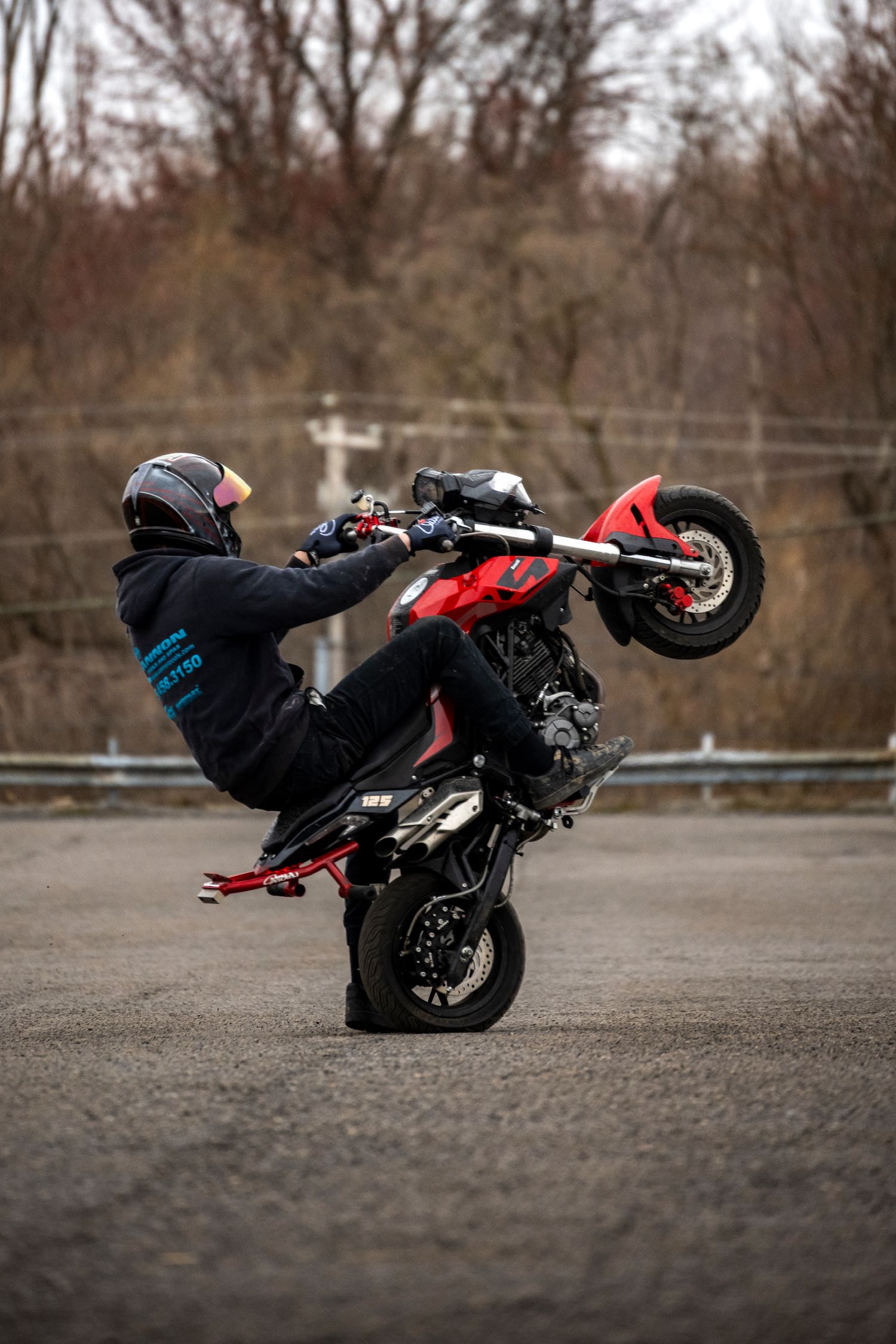 Stunt rider performing a wheelie on a red motorcycle in an empty parking lot with bare trees in the background.