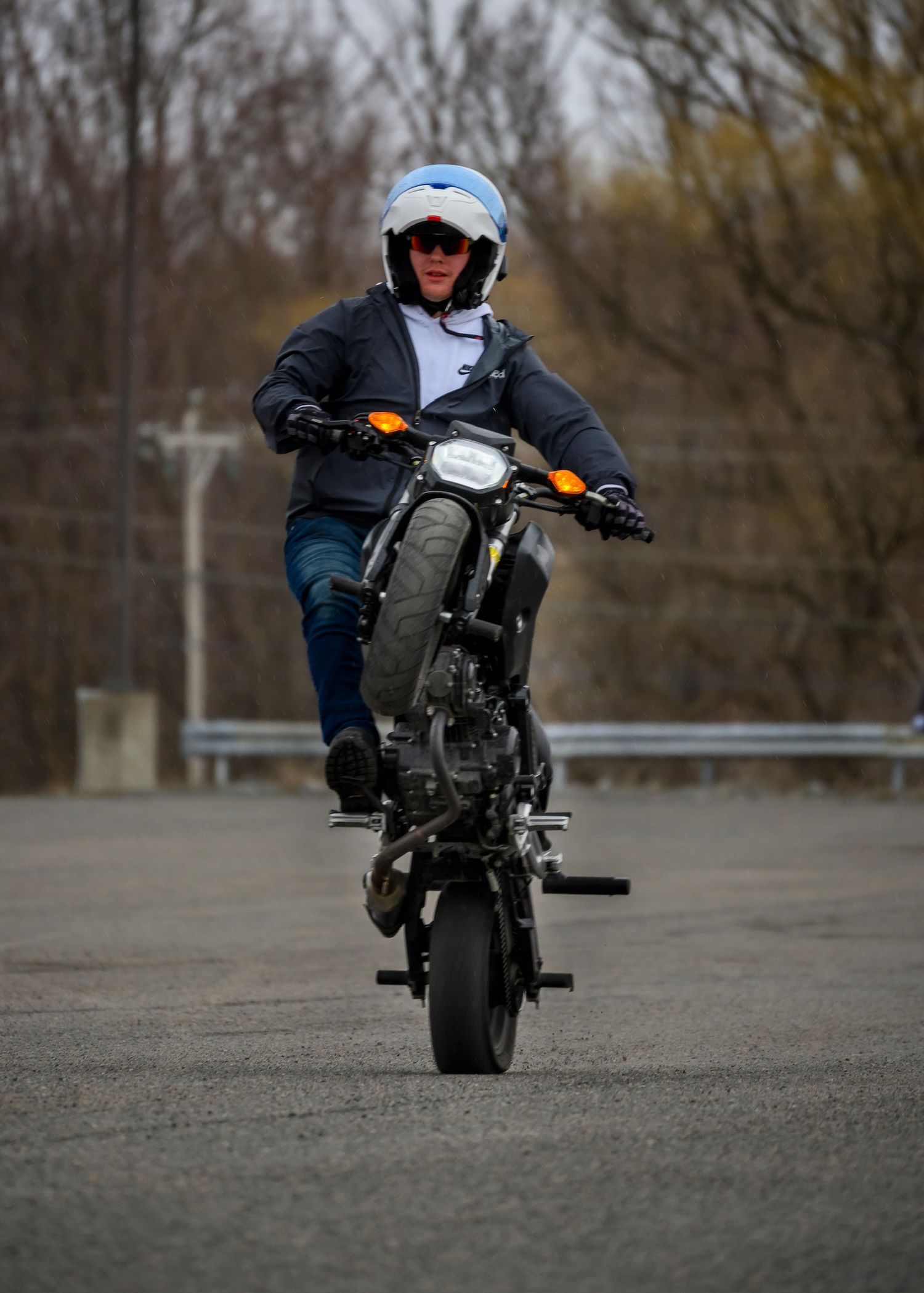 Motorcyclist performing a wheelie stunt on a dark motorcycle, wearing a blue helmet and jacket on an open pavement.