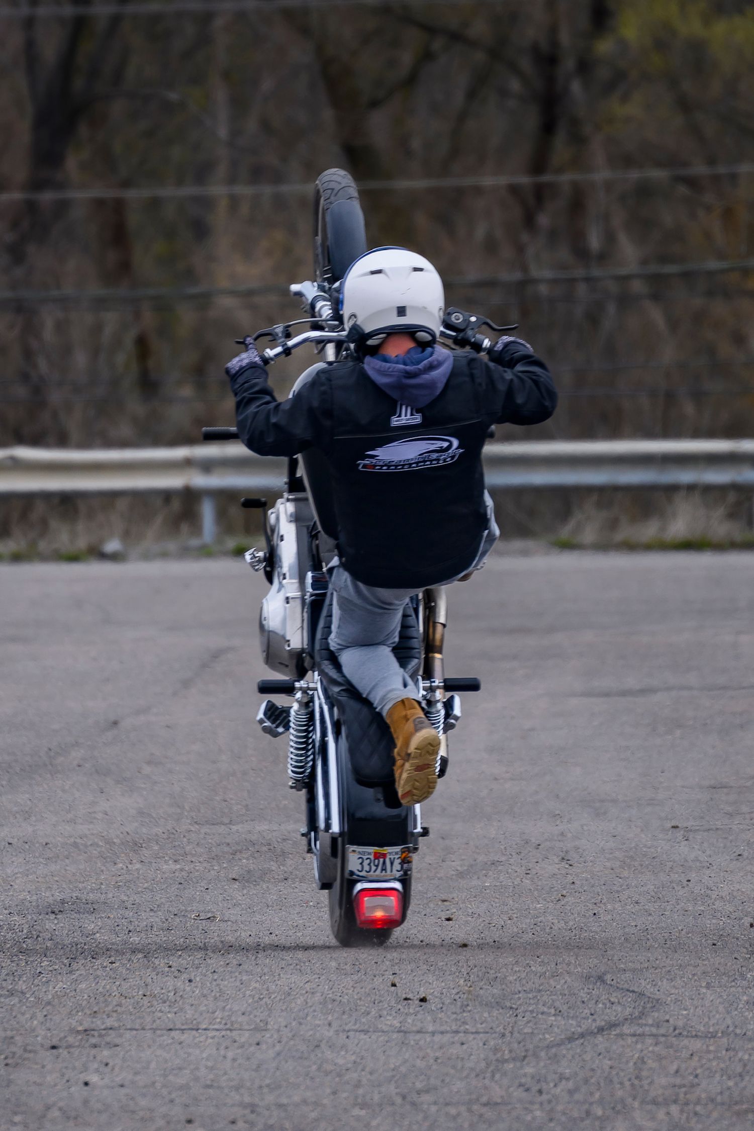 Motorcyclist performing a wheelie stunt on a silver bike, wearing a white helmet and black hoodie, on an empty road.