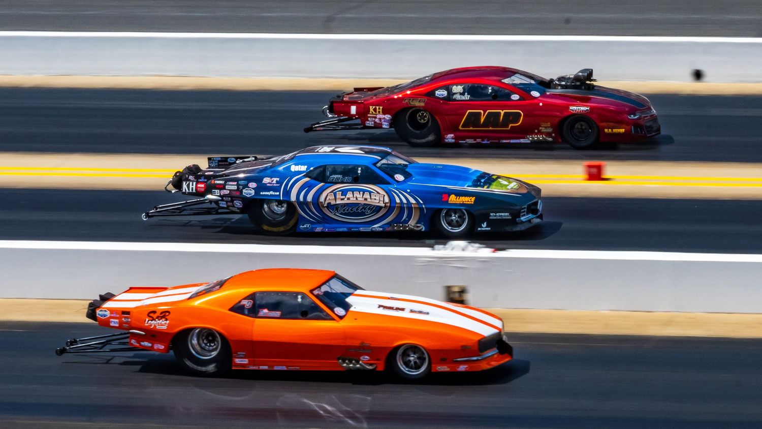 Colorful pro mod drag racing cars competing side by side on race track.
