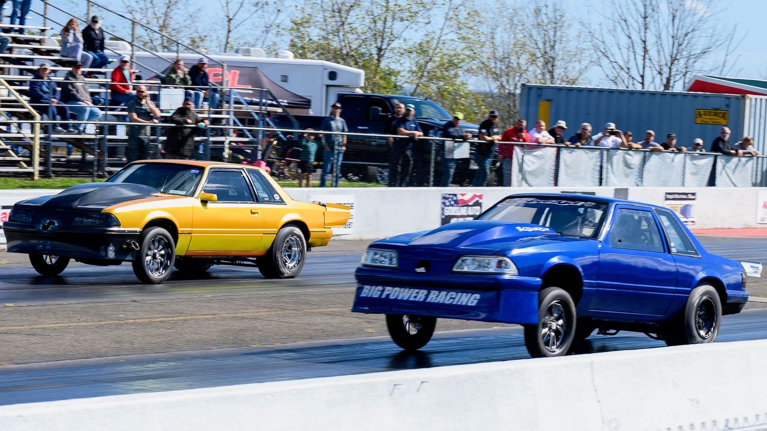 Classic muscle cars lined up at a drag racing event on a sunny afternoon.