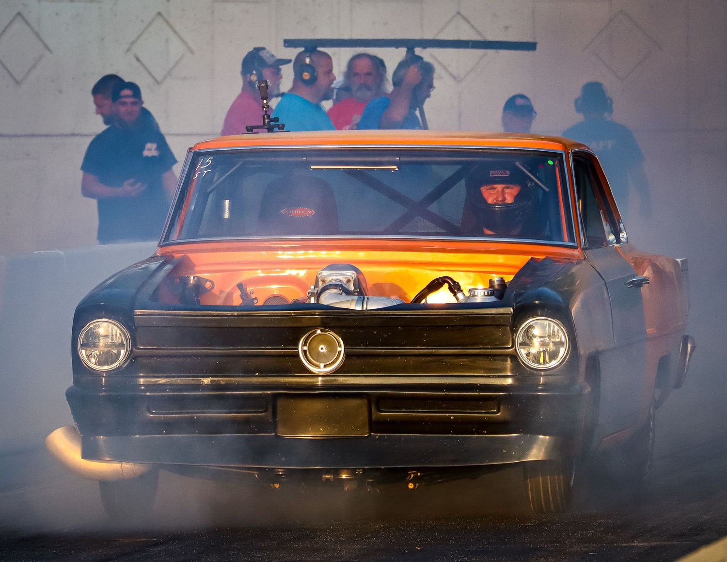 Classic Cadillac performs a smoky burnout at a drag racing event with spectators watching in the background.