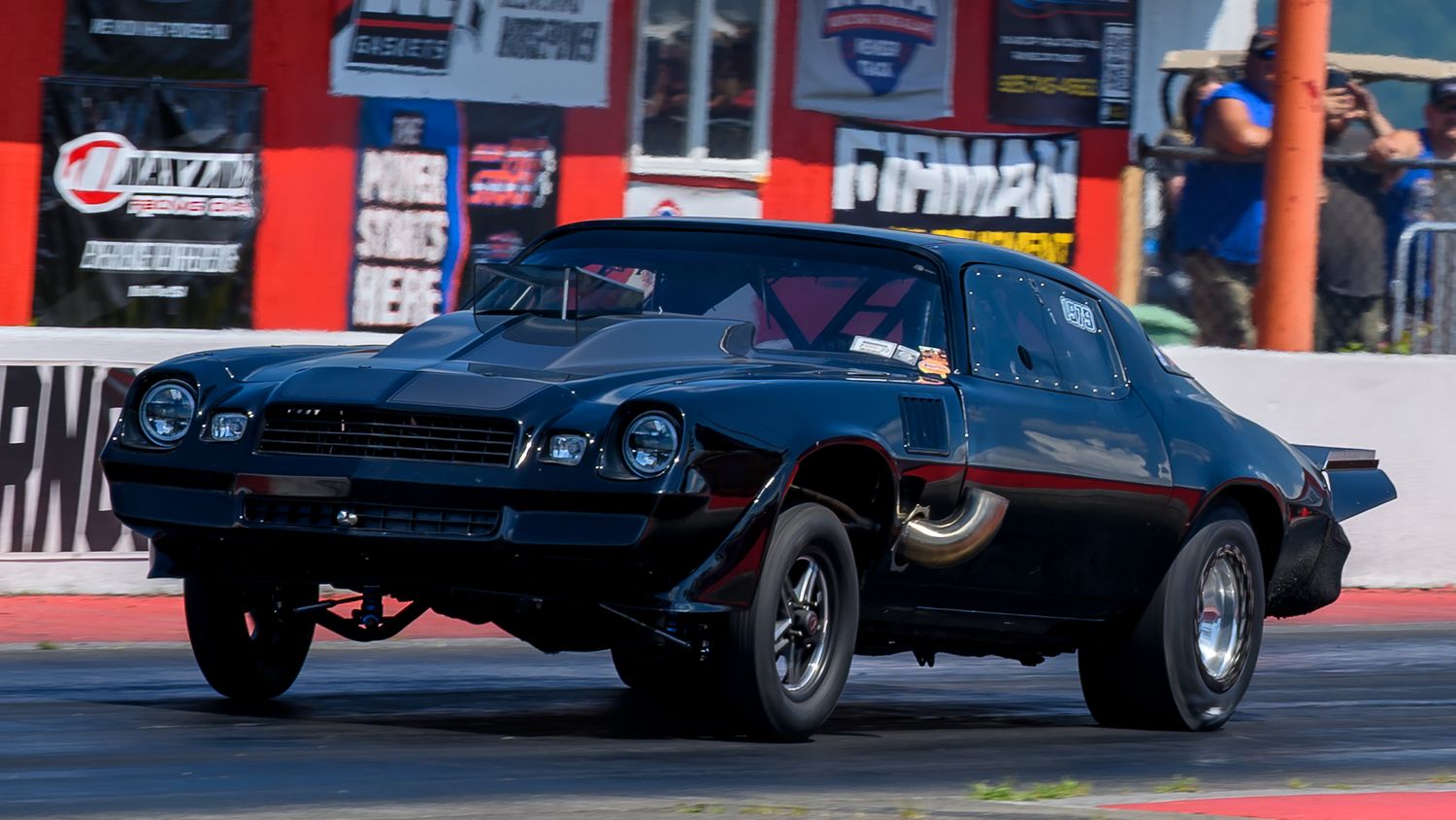 Dark drag racing cars perform on a track with red and white sponsor banners visible.