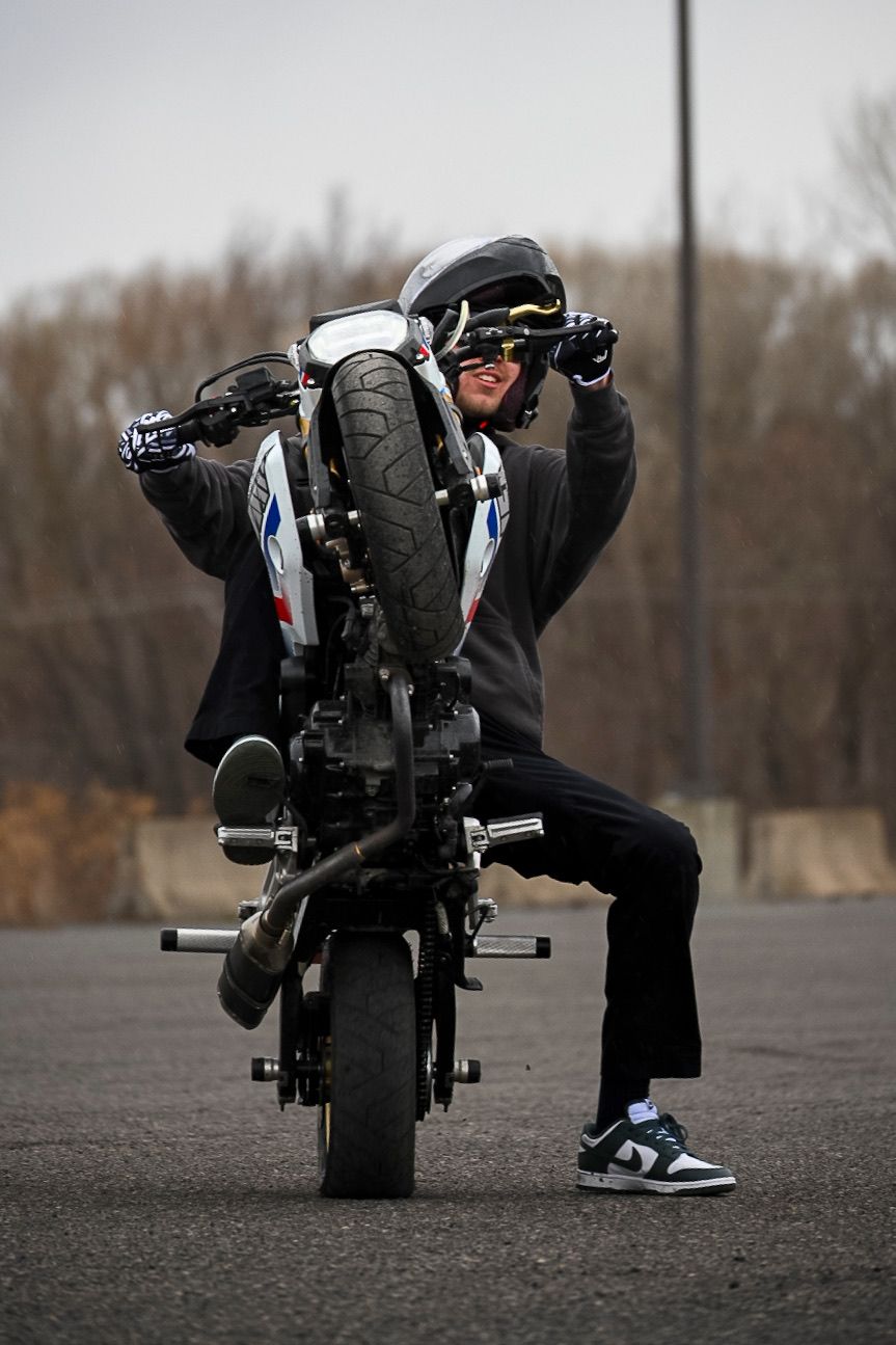Motorcyclist performing a stoppie stunt on a sport bike in a parking lot, wearing helmet, gloves, and Nike sneakers.