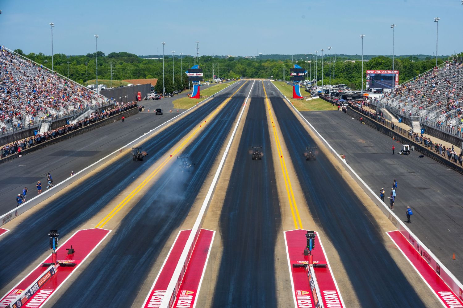 Wide angle view of a drag racing track with red lanes and grandstands on a sunny day.