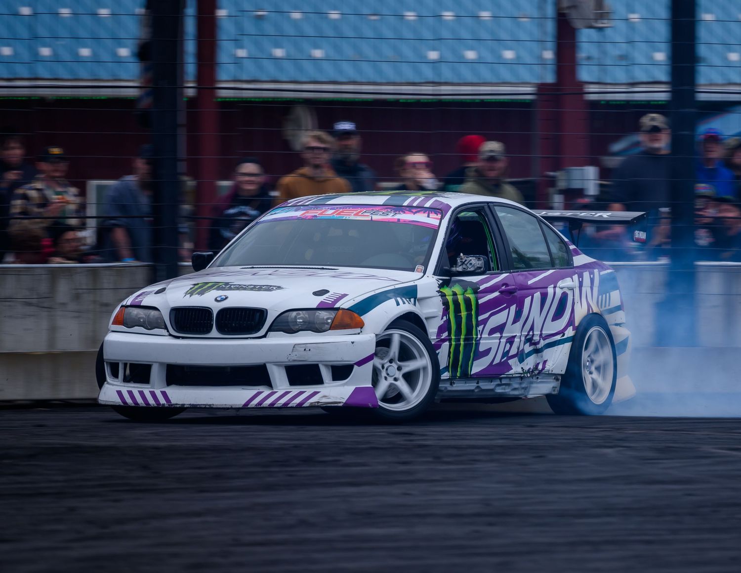 BMW drift car with Monster Energy sponsorship performing smoky drift at motorsport event with crowd watching.