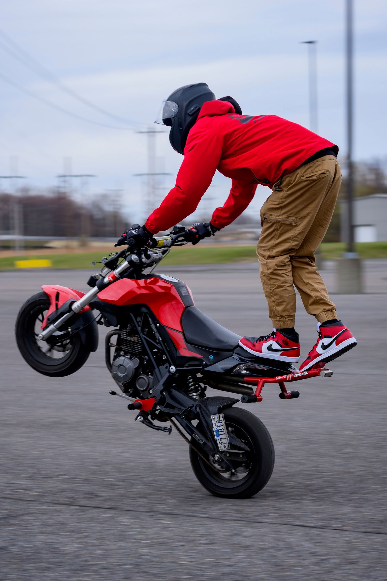 A stunt rider in a red hoodie and Air Jordans performs a wheelie on a red motorcycle in an open parking lot.
